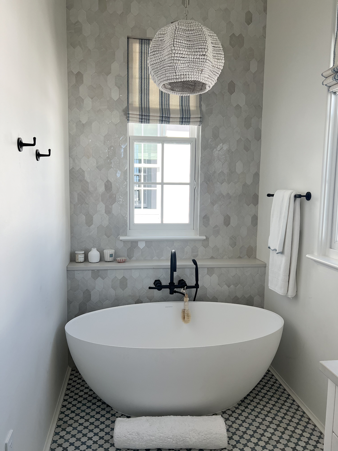 A modern bathroom with a white freestanding bathtub, black faucet, and a window with a striped valance. The wall behind the tub features hexagonal tiles, and there are black hooks and a towel bar. A white rug is on the floor.