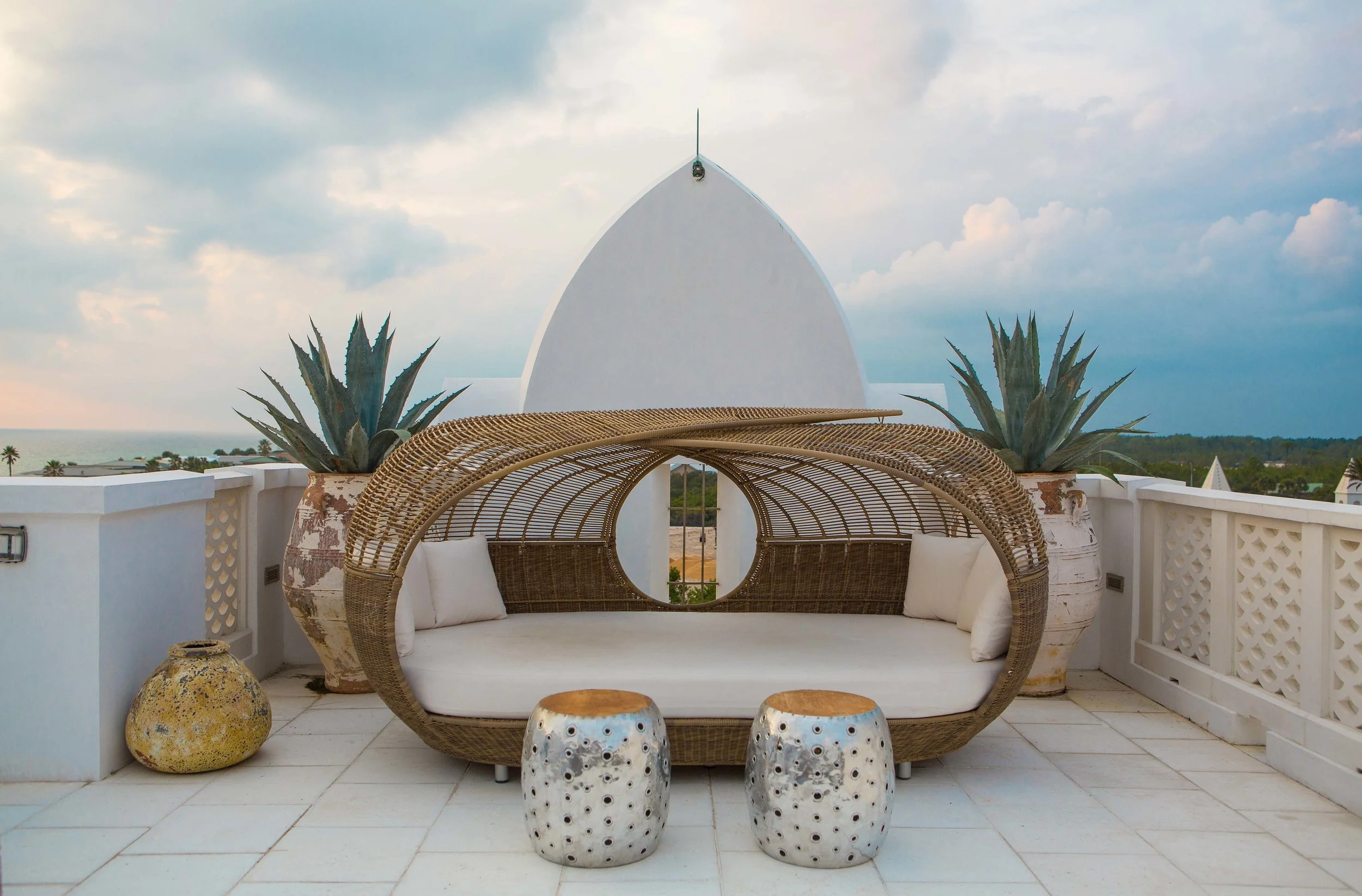 Outdoor seating area on a rooftop with a bamboo and rattan daybed, two white ceramic stools, large potted agave plants, and a scenic ocean view sky in the background.