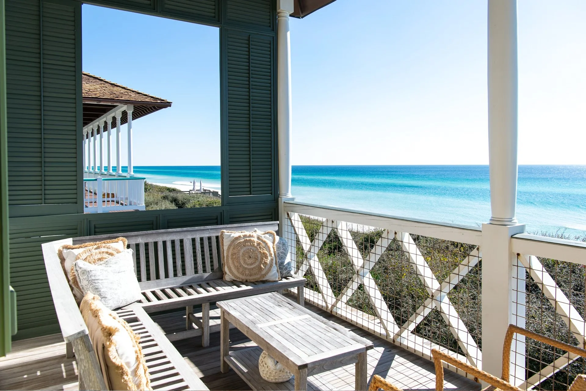 Beach view from a balcony with wooden furniture, cushions, and white railings, overlooking a sandy shoreline and ocean waves.