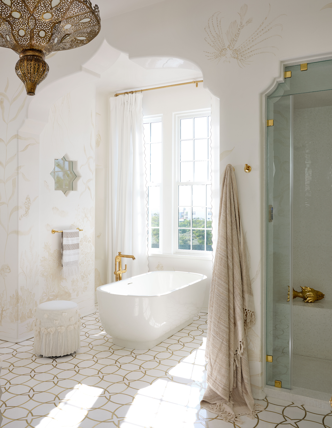 Bright and elegant bathroom with a white freestanding bathtub, large windows, gold fixtures, patterned floor tiles, a small upholstered stool, and a glass shower with gold details. Decor includes a wall mirror and soft neutral towels.