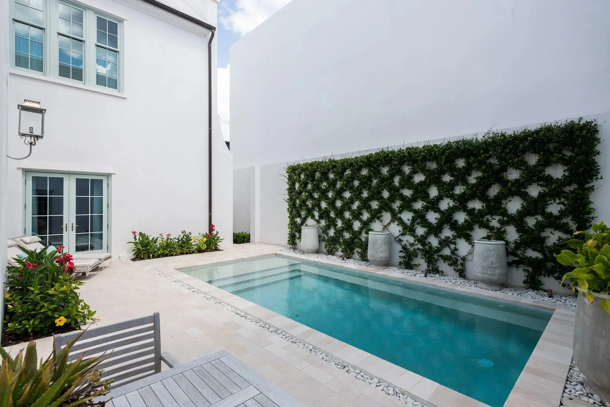 Small outdoor swimming pool surrounded by white stone tiles, with potted plants and flowers, a white wall with climbing greenery, and a modern white house with large windows in the background.