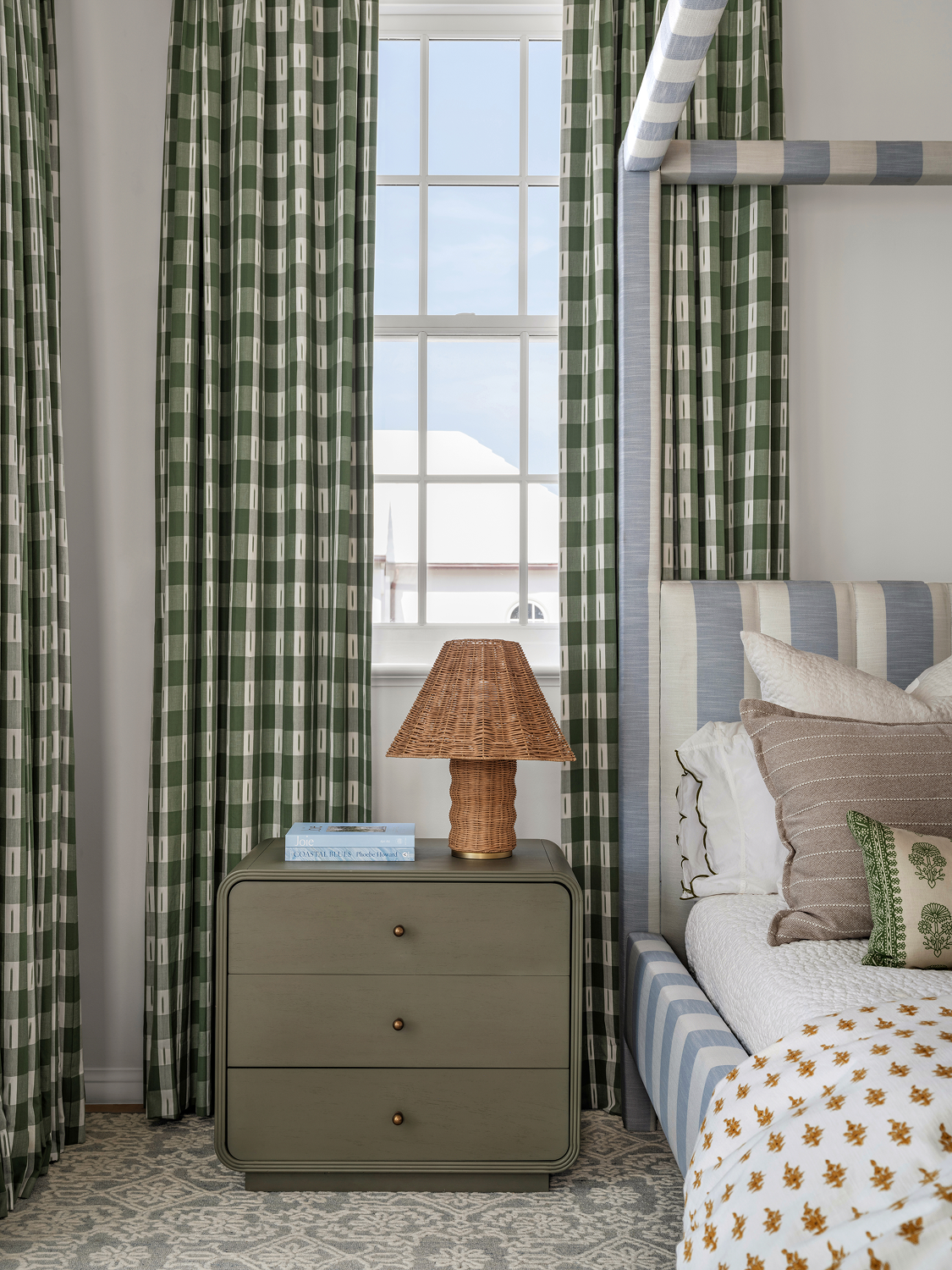 A bedroom with green plaid curtains, a window showing a white building outside, a three-drawer bedside table with a wicker lamp, some magazines, a bed with pillows, and a blue-striped headboard.