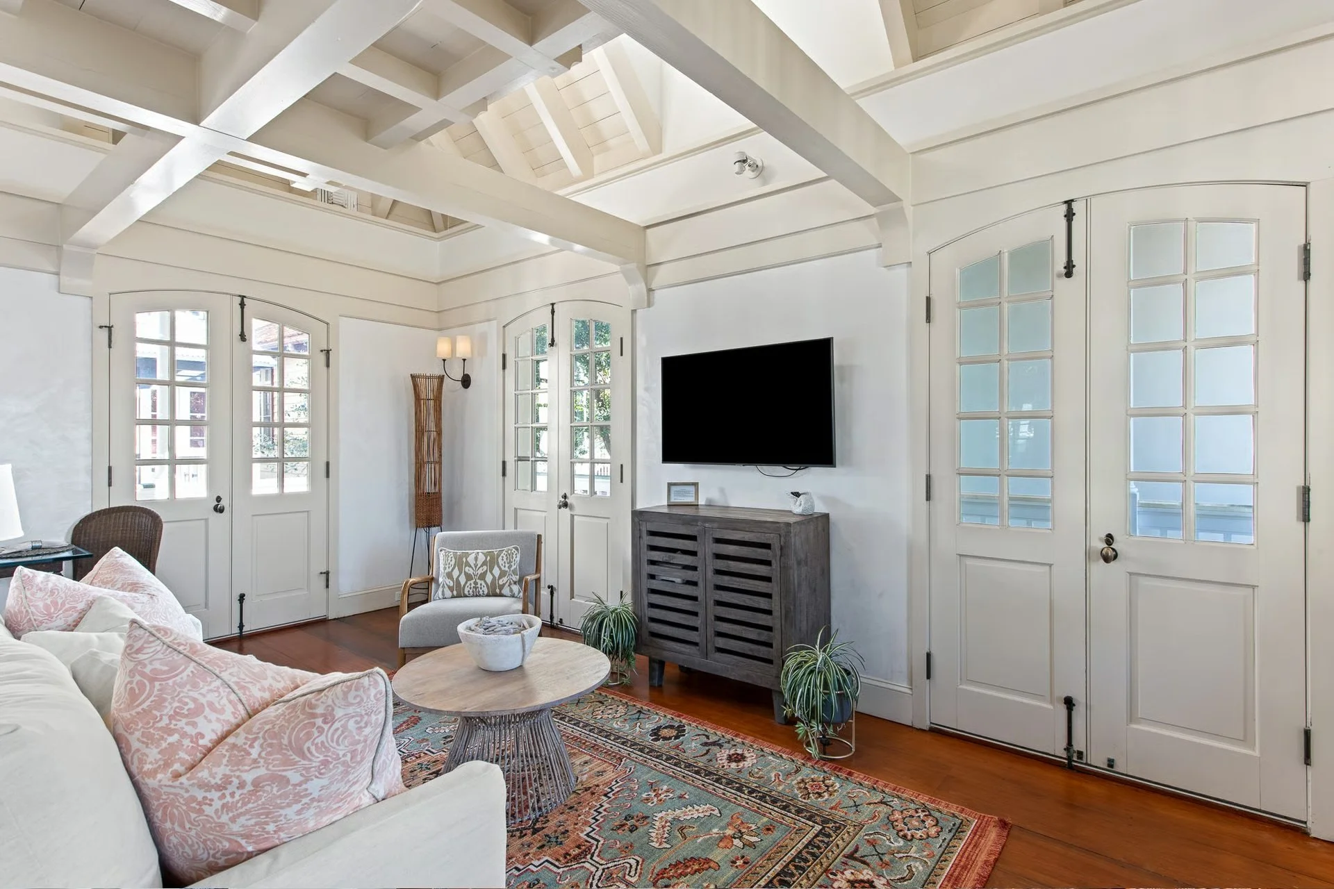 Living room with white walls, three glass-paneled doors, a wall-mounted TV, a gray cabinet, a beige sofa with pink and white pillows, a wooden coffee table, a patterned area rug, and green plants.