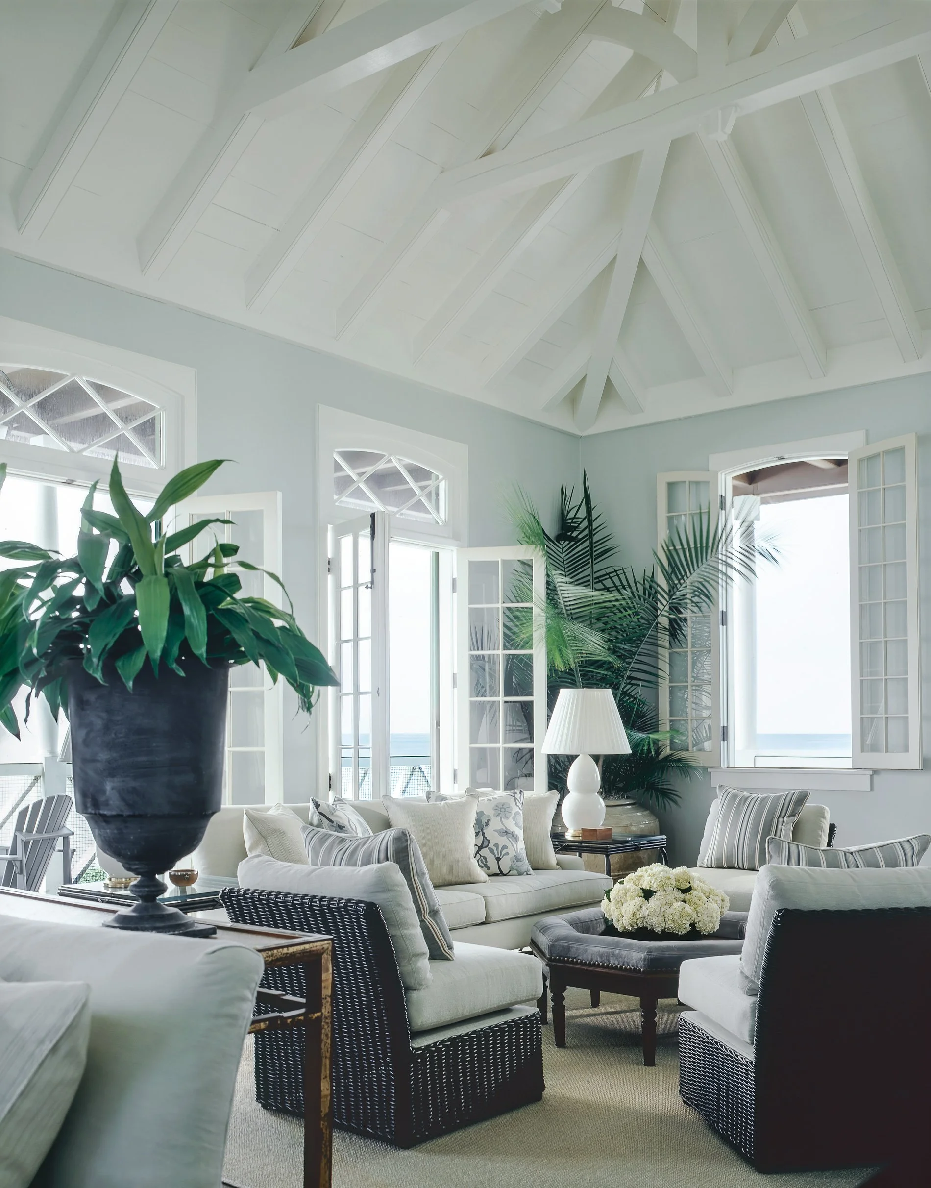 Bright living room with open windows, white furniture, large plants, and a high vaulted ceiling.