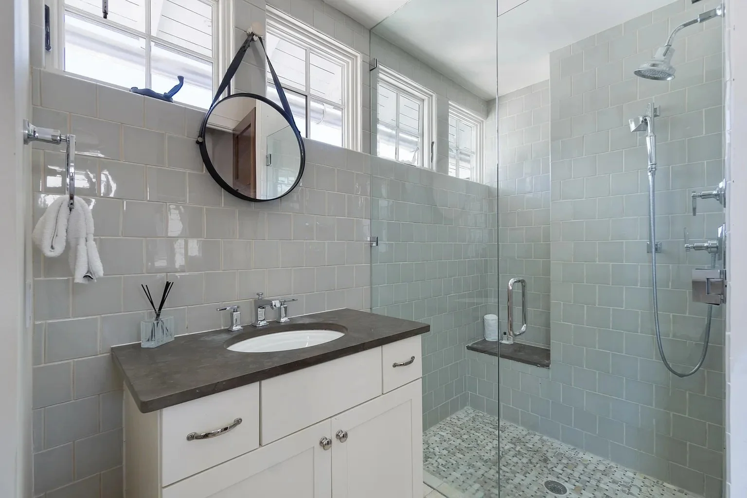 Modern bathroom with a white vanity, dark countertop, oval sink, wall mirror, towel rack with towels, glass-enclosed shower, tiled walls, and windows above the shower.
