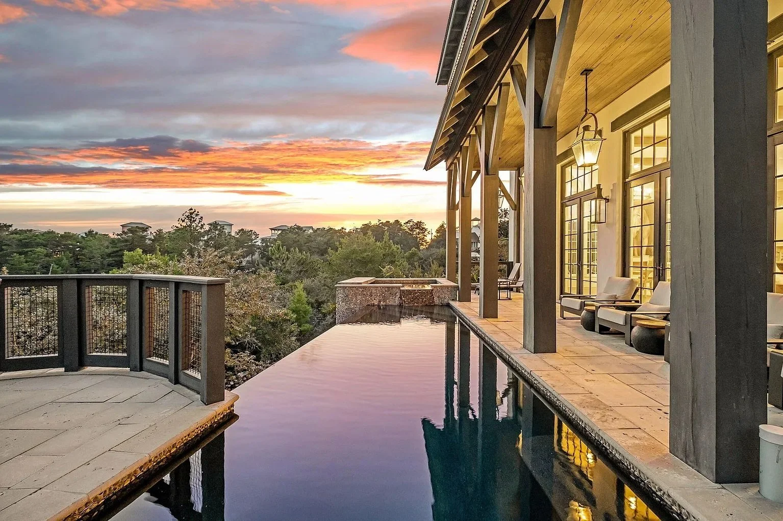 An outdoor swimming pool overlooking a sunset with a wooden deck, railing, and outdoor seating area with chairs and wall-mounted lamps.