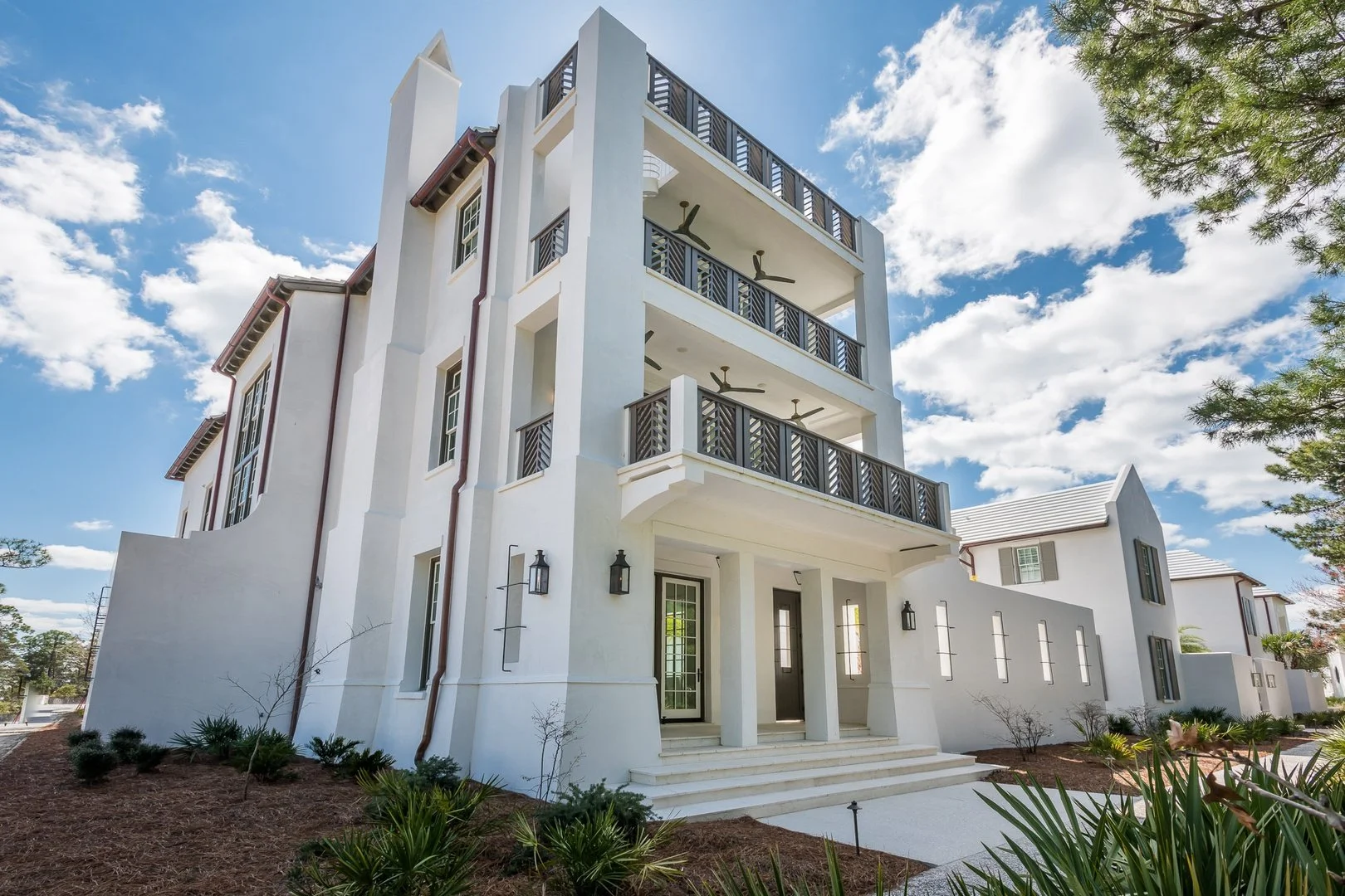 A modern white multi-story apartment building with balconies, black railings, and ceiling fans, set against a partly cloudy blue sky with some trees and landscaped ground in the foreground.