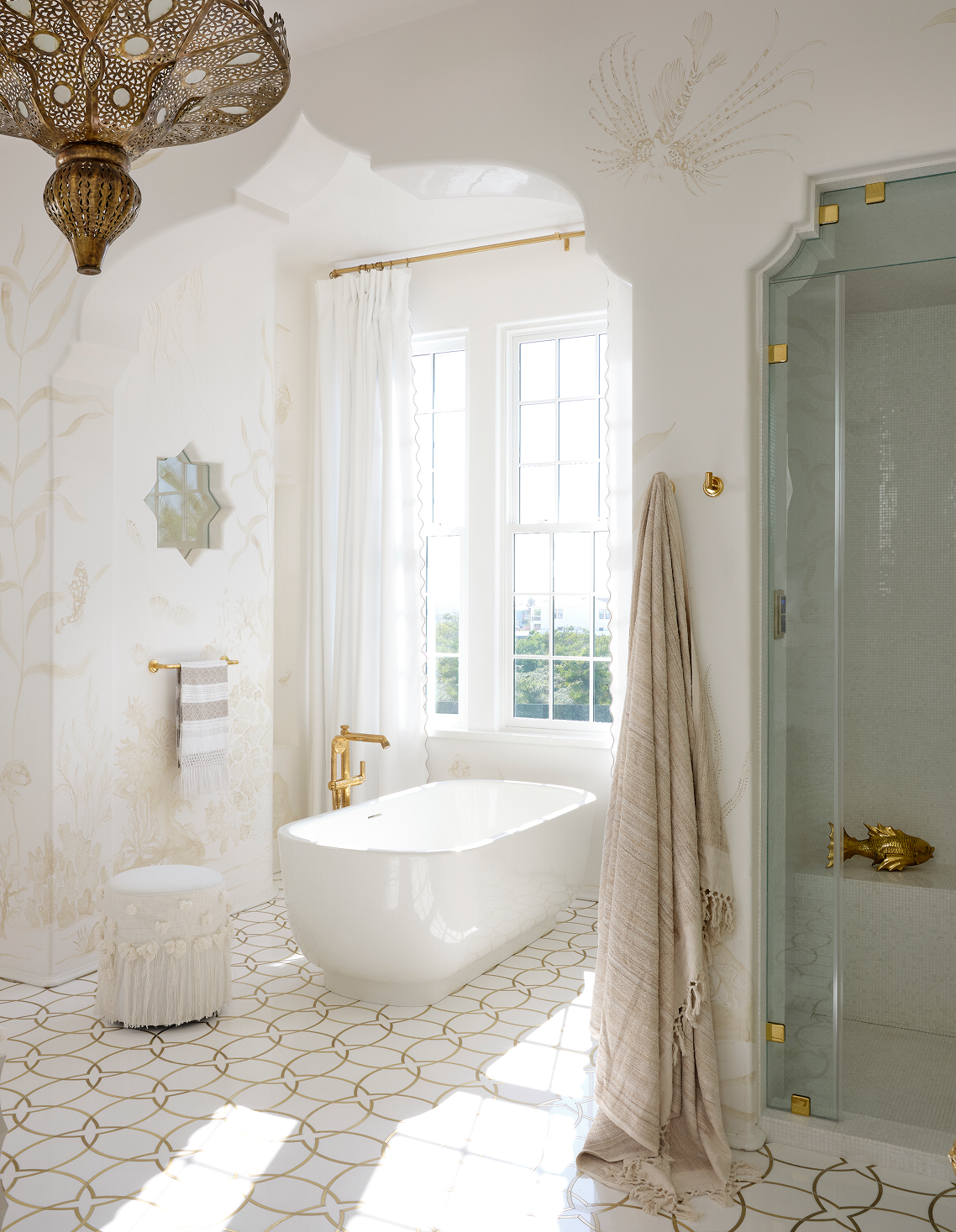 Bright bathroom featuring a modern freestanding bathtub with gold fixtures centered under two large windows, a glass shower with gold accents, ornate gold and woven light fixture, beige towels hanging on the wall, a small round white stool with fring