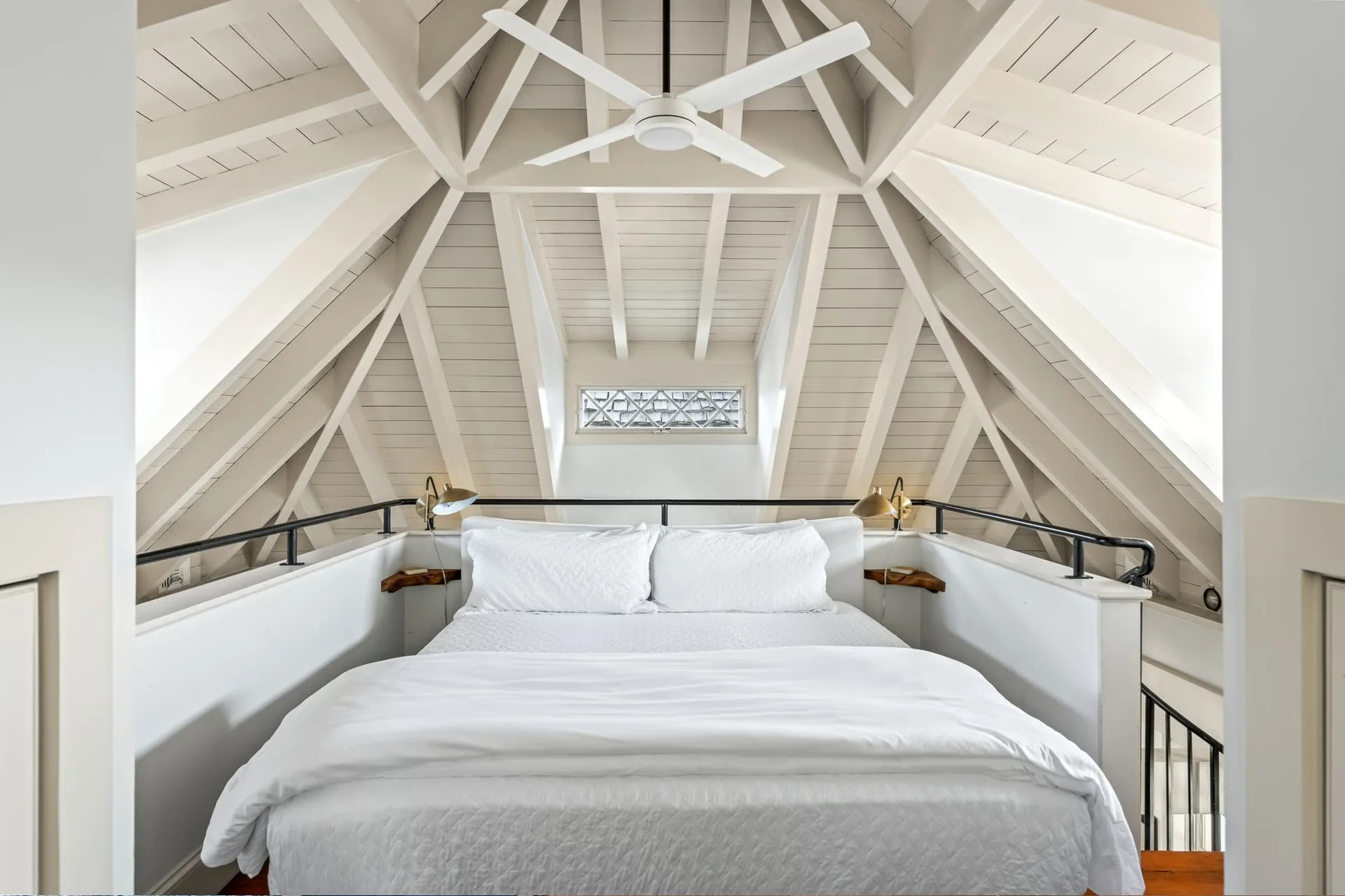 A cozy bedroom with a white bed, wooden side tables, brass wall lamps, and a white ceiling fan, situated in an attic space with sloped white-paneled ceiling beams.