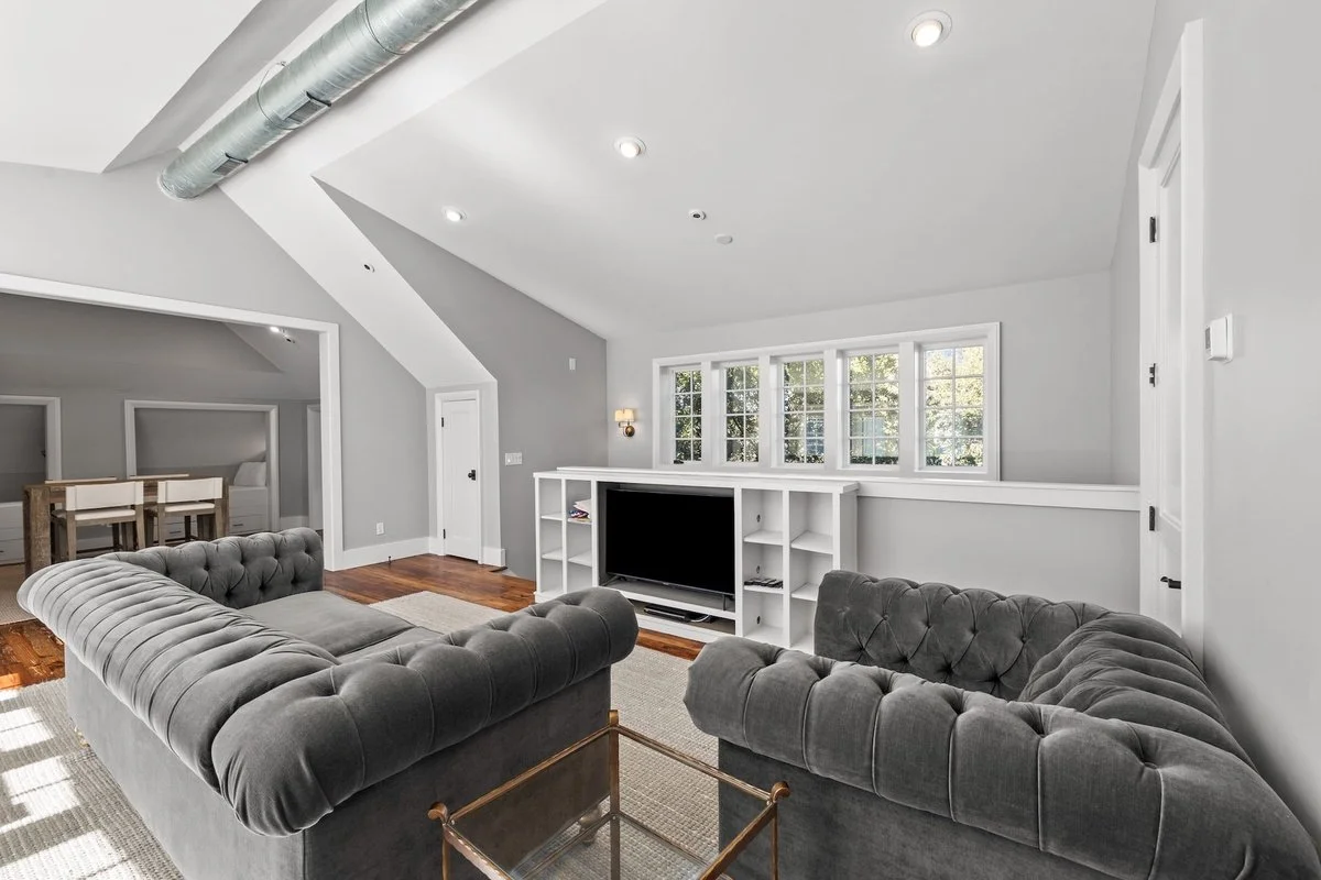 Living room with two gray tufted sofas, a white TV stand with a large TV, and a set of three windows letting in natural light. Hardwood floors and gray walls complete the space.