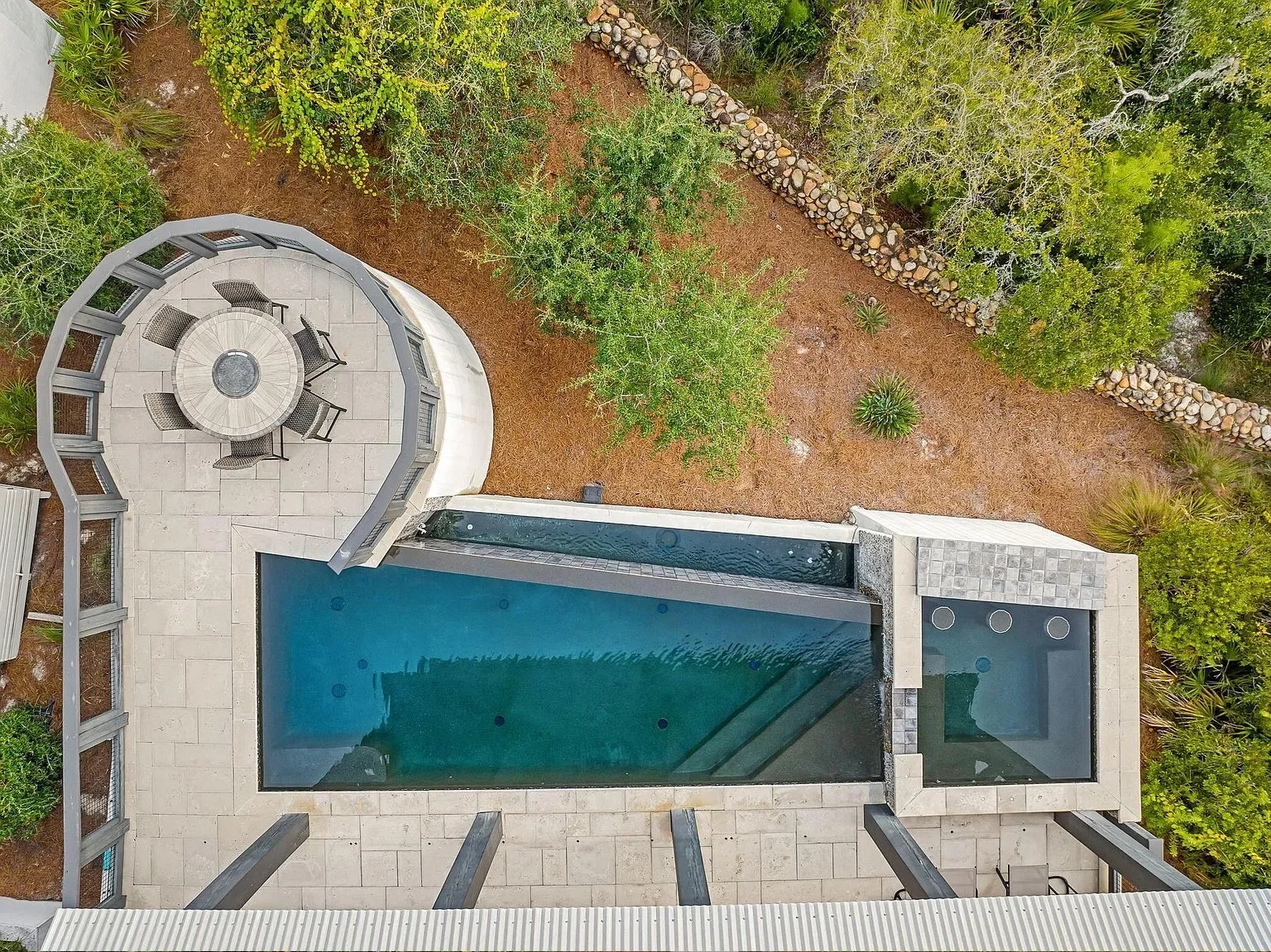 Aerial view of a backyard with a swimming pool, hot tub, outdoor dining area with chairs and a round table, surrounded by trees and a stone wall.