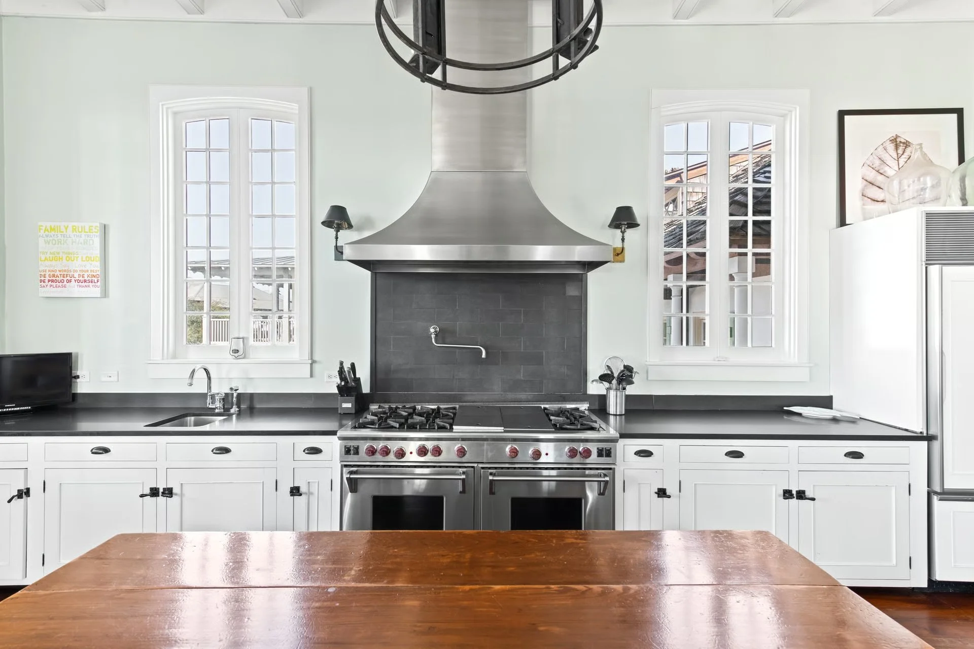Modern kitchen with white cabinets, black countertops, and stainless steel stove with a black range hood. Two windows above the countertop, and decorative items including a framed art piece and a refrigerator.