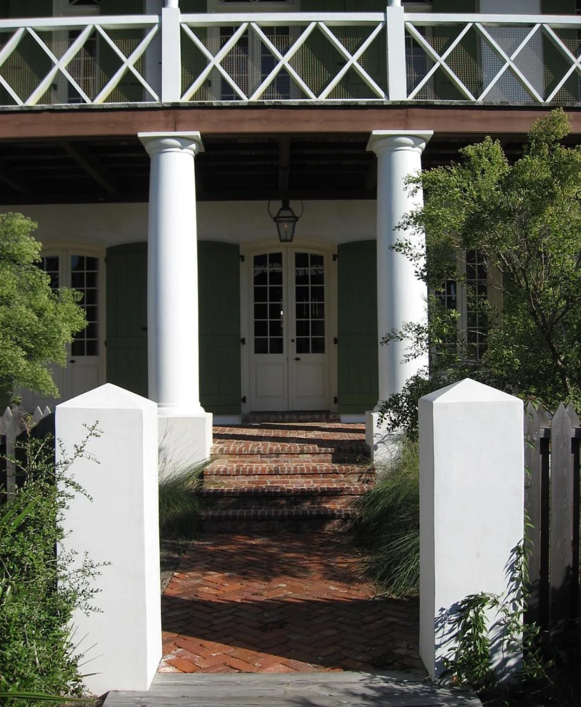 Front view of a house with a brick pathway, white posts, green shutters, a balcony, and trees.