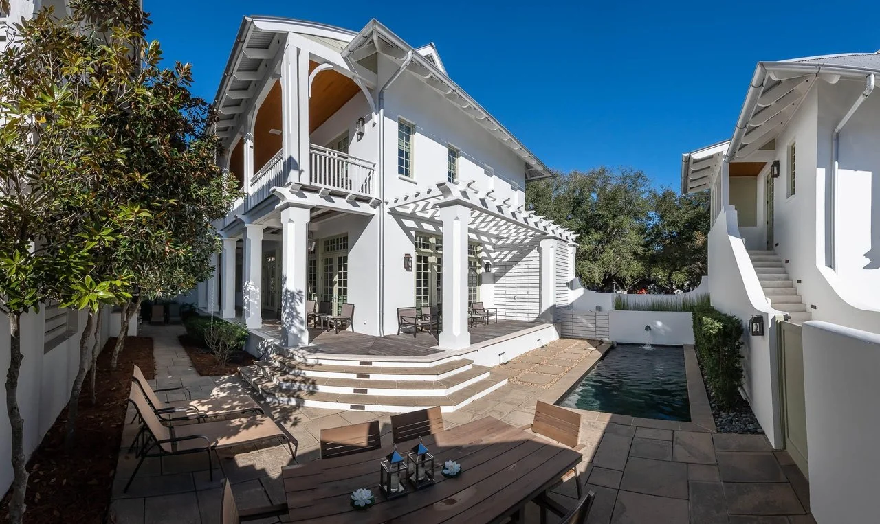 A modern white two-story house with large windows and a balcony, surrounded by a backyard with a patio, outdoor furniture, and a small swimming pool. There are trees and plants around the yard, and clear blue sky overhead.