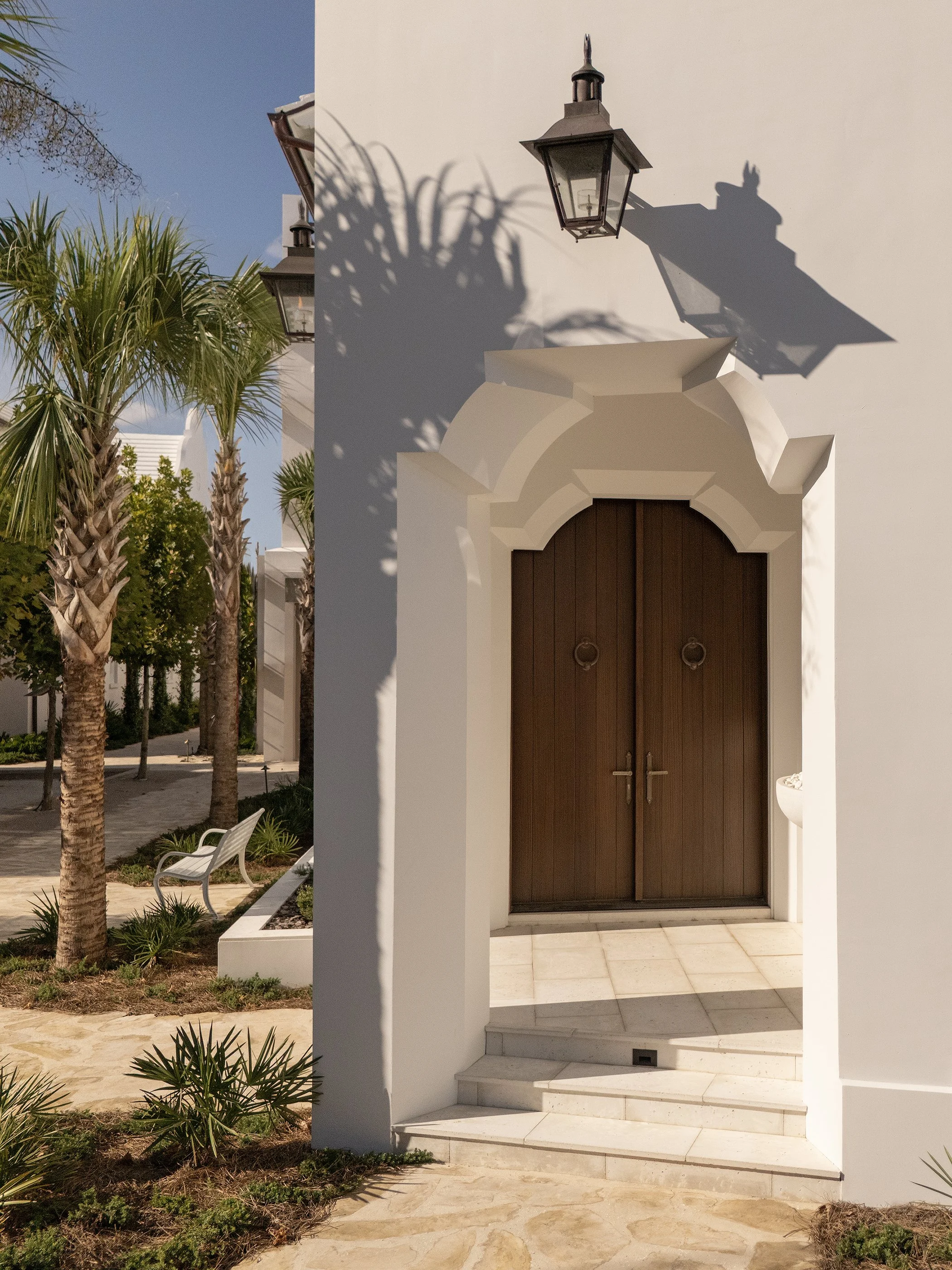 White exterior wall with a dark brown wooden door and decorative archway, shadows of a lantern and plant on the wall, and an outdoor garden area with palm trees and a bench.