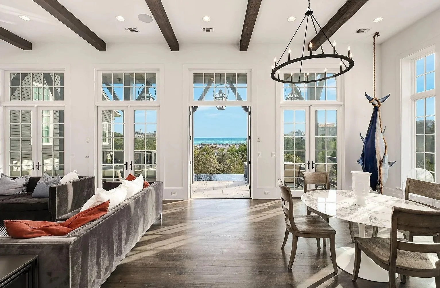 Living room with large windows and a door leading to a balcony, view of the beach and ocean, modern decor, chandelier, sofa with cushions, and a dining table with chairs.