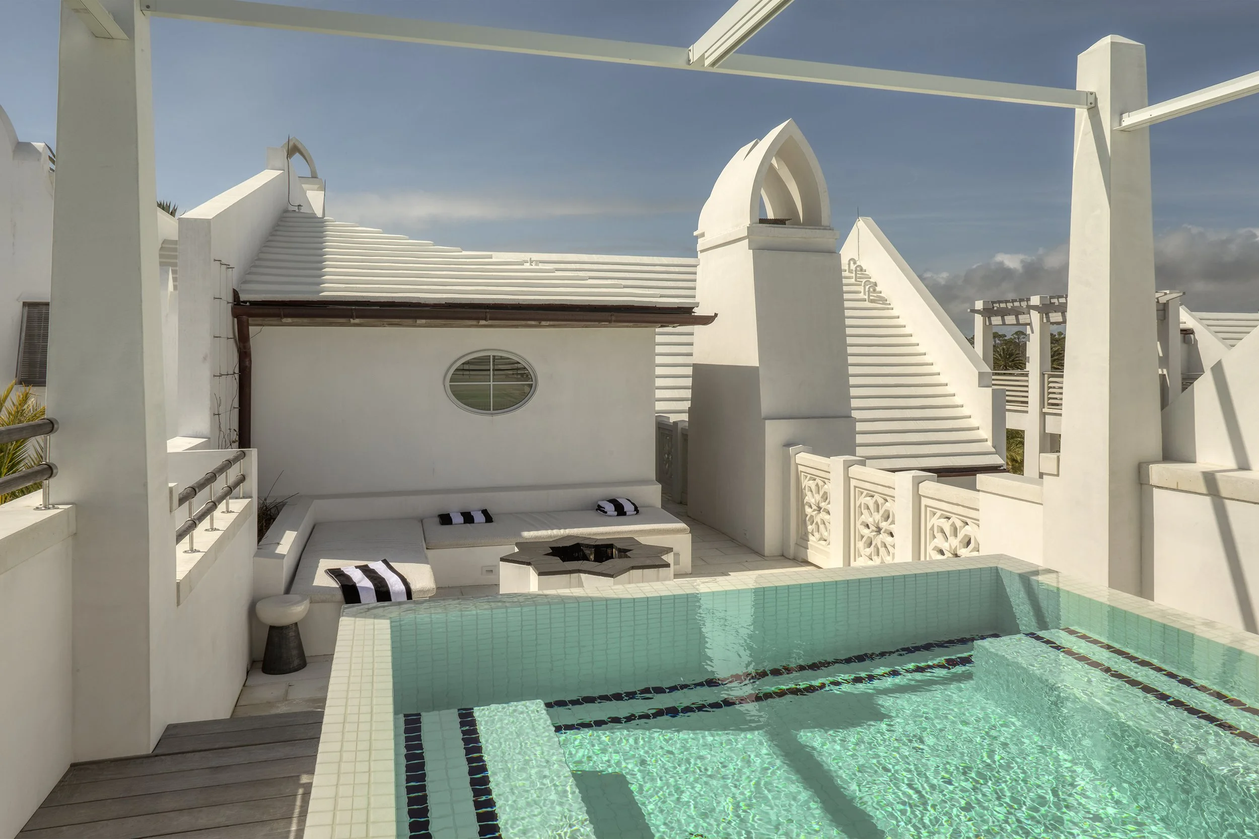 A modern white building with stairs leading to an upper level, round window on the wall, a swimming pool in front, and outdoor seating with striped cushions.