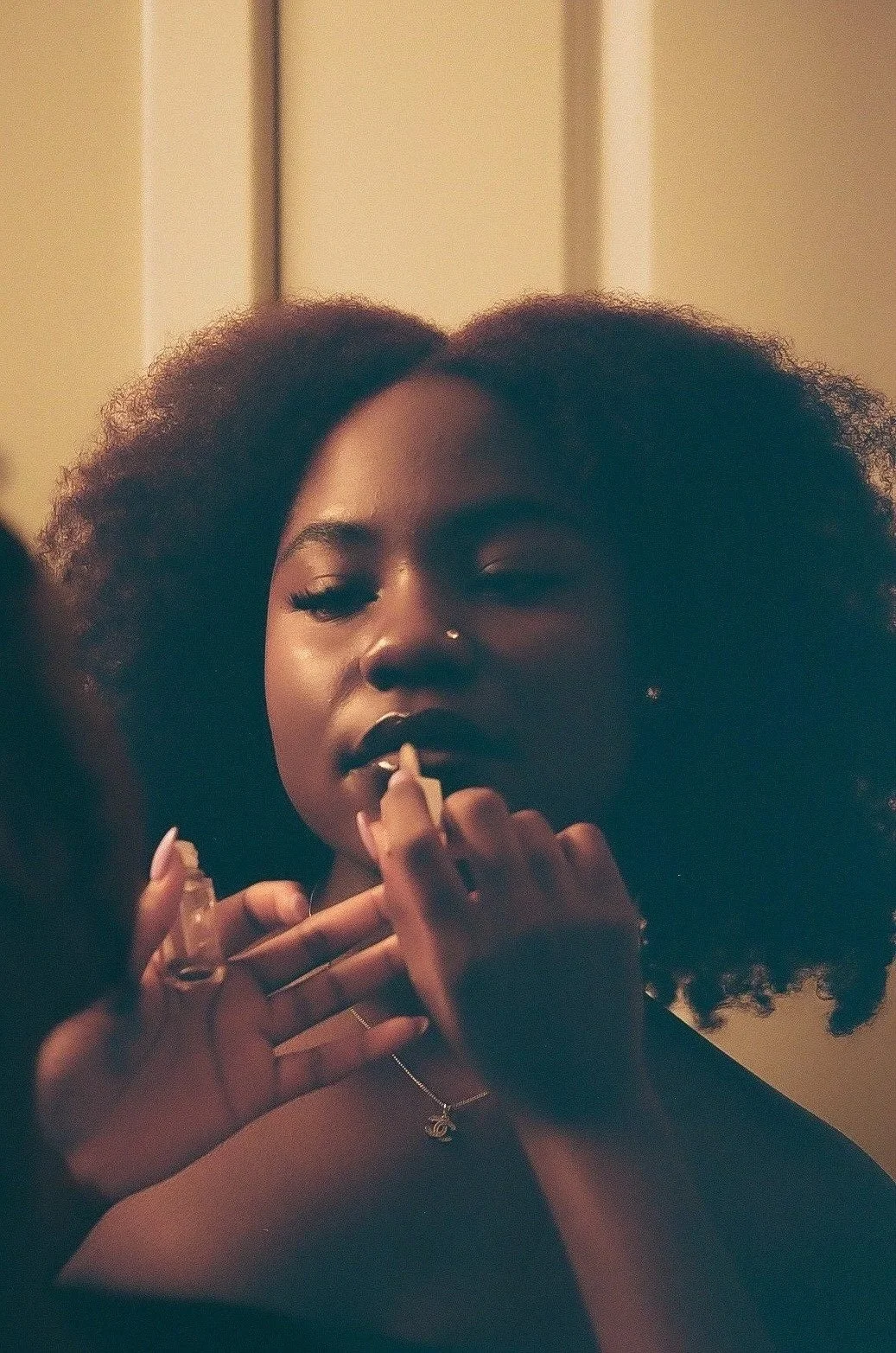 A woman with curly hair applying lipstick in front of a mirror, wearing a necklace and a dark top.