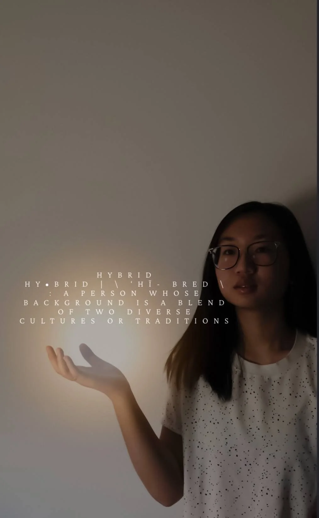 A woman with glasses wearing a white shirt with small black dots, standing in front of a plain wall with text about hybrid culture, holding her hand up as if presenting.