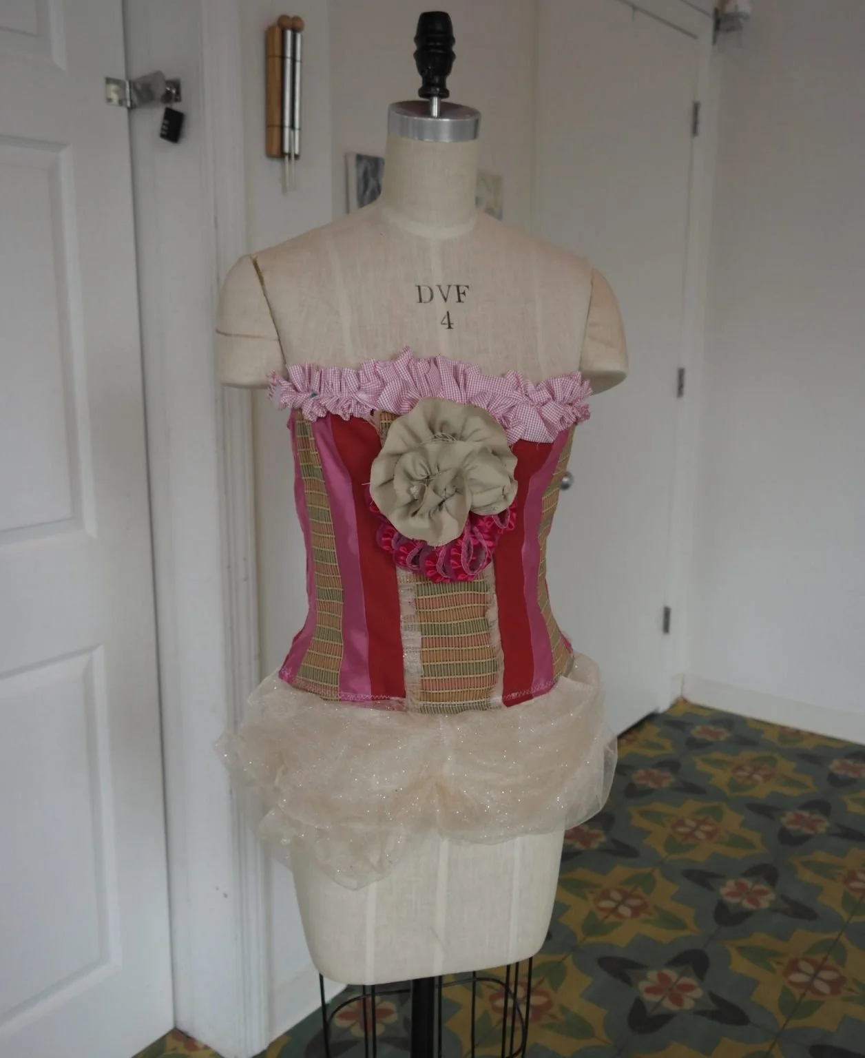 A mannequin dressed in a colorful, striped corset with a pink ruffled top edge and large fabric flower in the center, standing indoors against a white door and wall with patterned carpet.