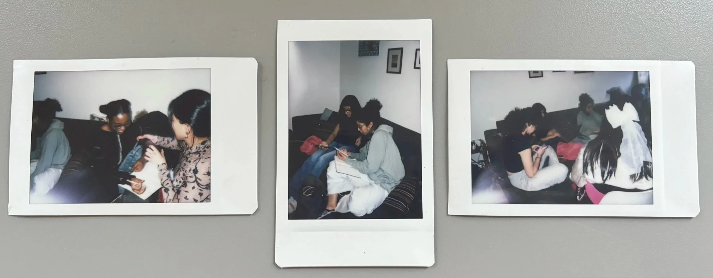 Three printed photographs of four young women sitting on a couch, engaged with their phones or talking, displayed on a gray wall.