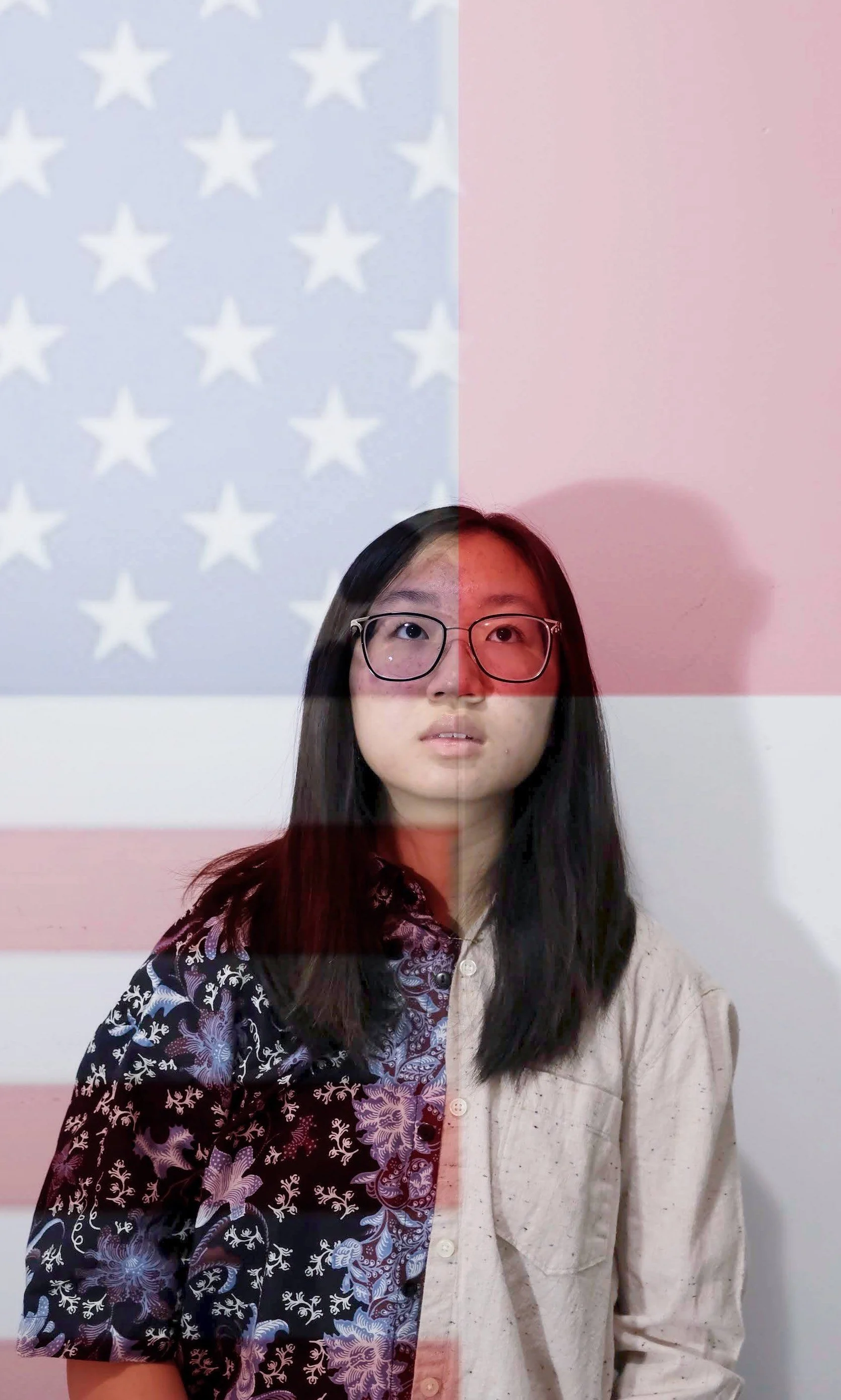A young woman with glasses and long black hair standing in front of a wall with a projection of the American flag.