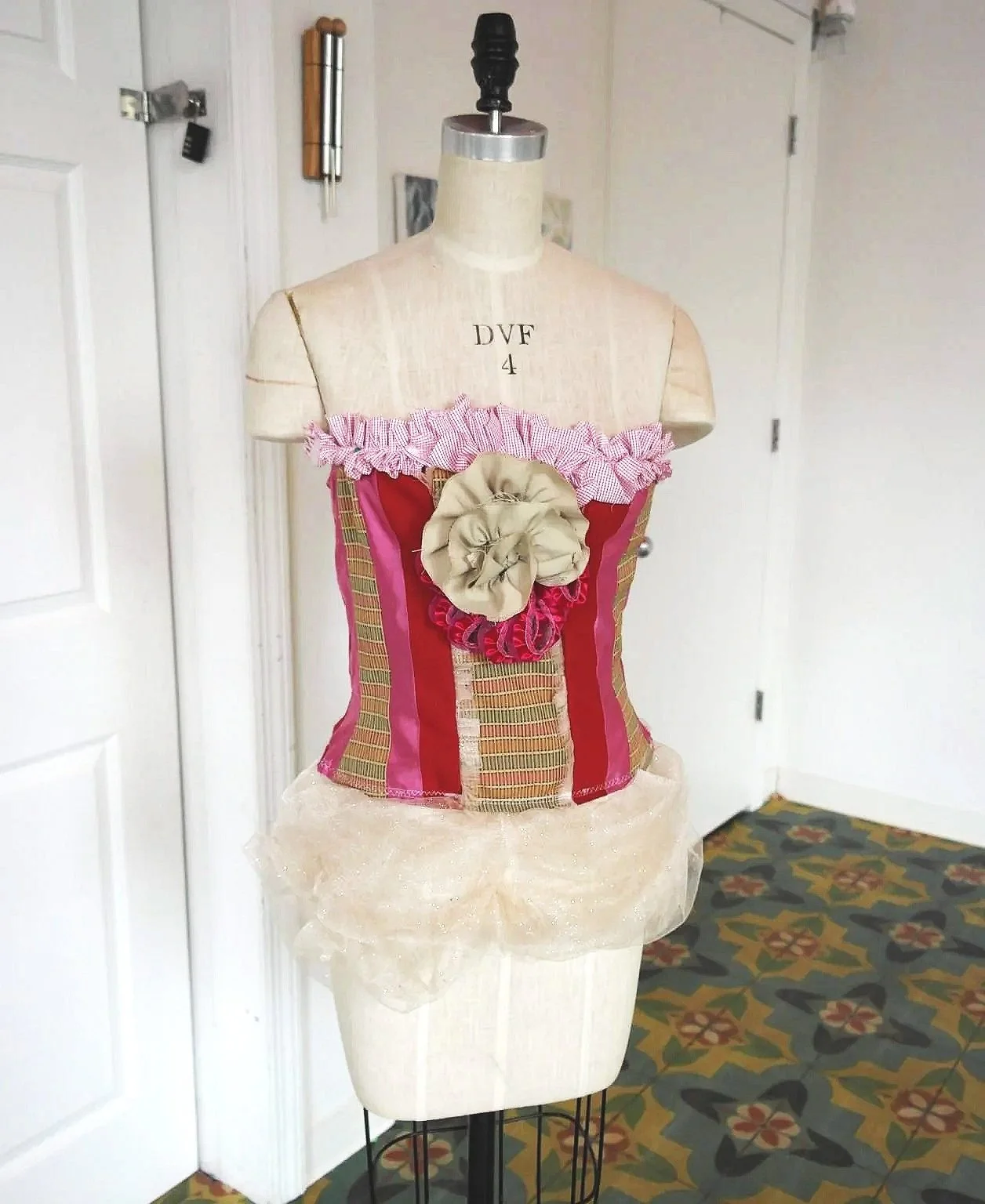 A dress form features a patchwork-style corset with red, beige, and patterned fabric, adorned with fabric flowers and pink and red accents, and a sheer, ruffled beige fabric at the bottom.