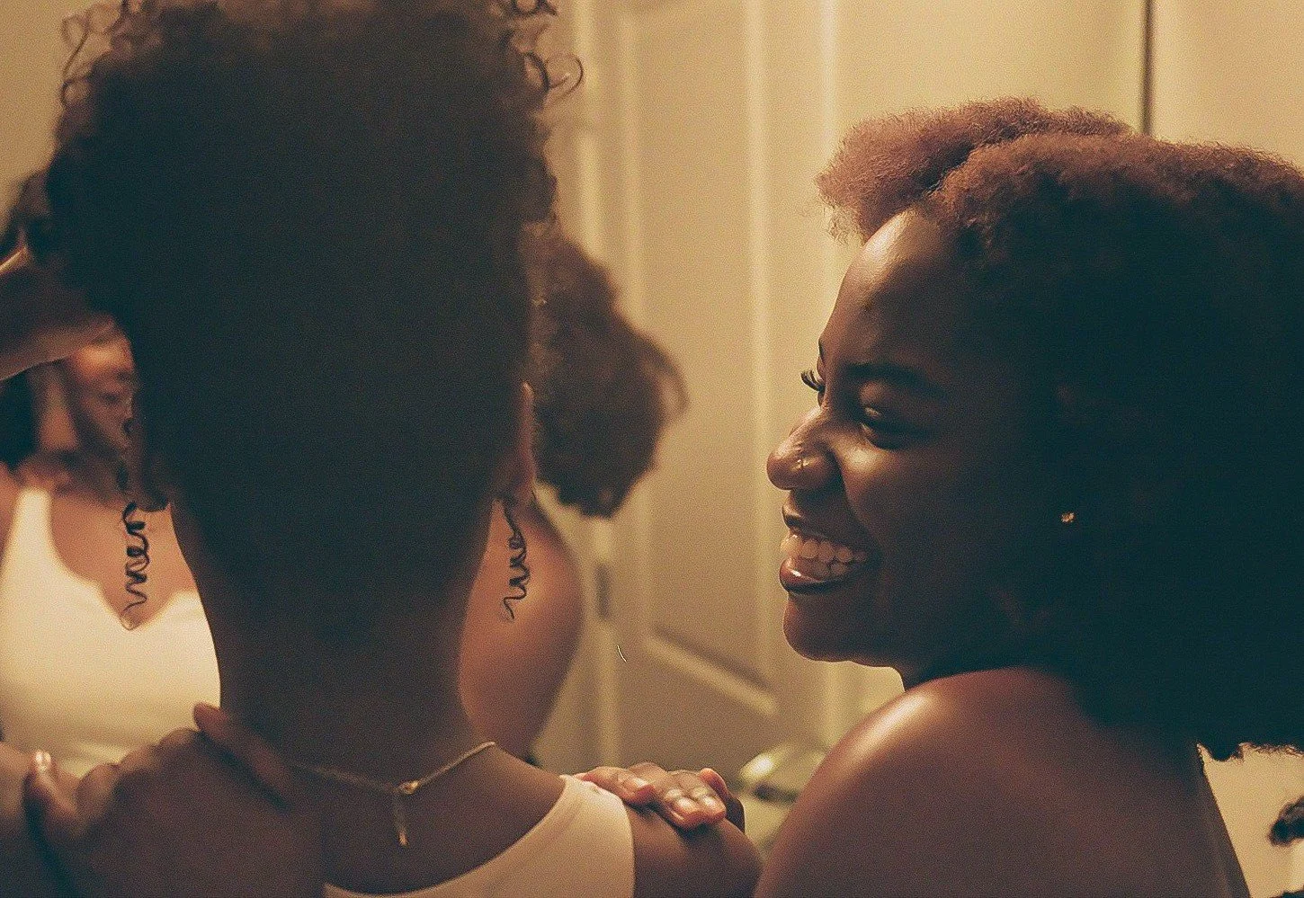 Two smiling women with natural hair sharing a moment in a warmly lit room.