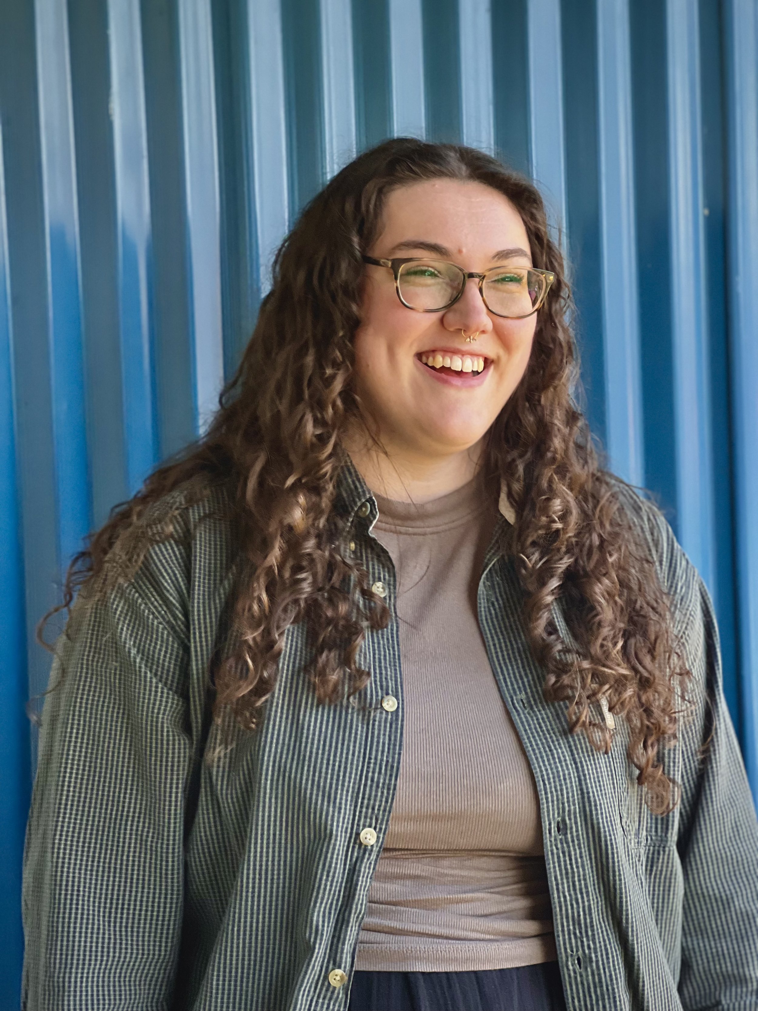 A white woman with long curly brown hair wearing a brown shirt under a green button-up shirt and glasses smiling in front of a blue background