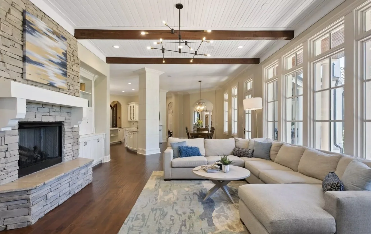 Living room with large sectional sofa, stone fireplace, wooden ceiling beams, modern chandelier, and large windows with a view outside, decorated with a rug, pillows, and a coffee table.