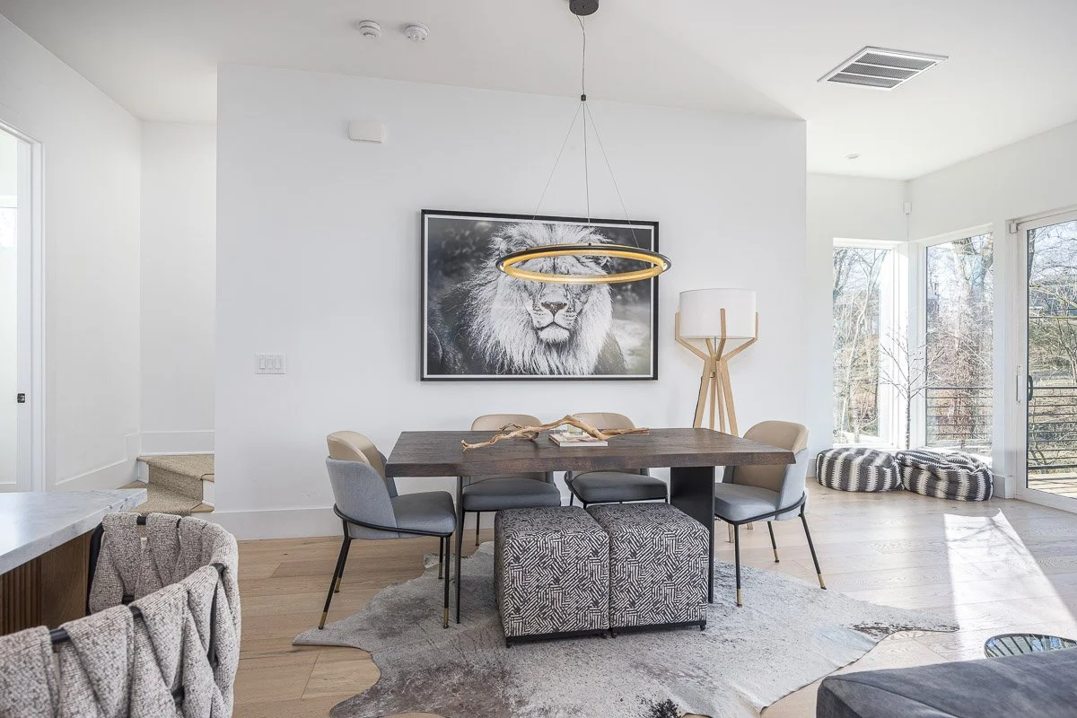 Modern dining room with a dark wooden table, six gray chairs, a black and white lion framed picture on the white wall, a unique tall tripod-style floor lamp, and two striped floor cushions near large glass sliding doors with an outdoor balcony view.