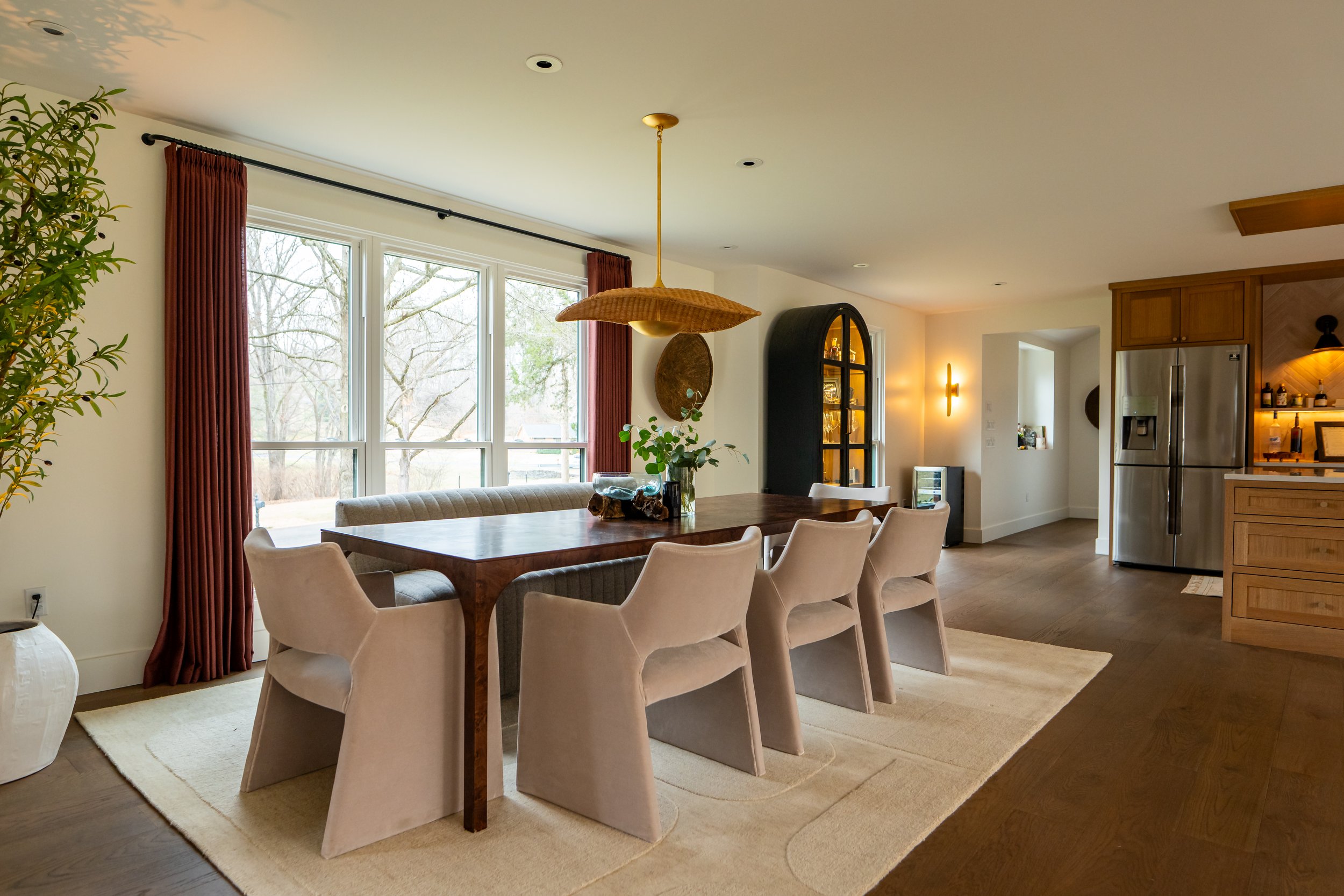 Modern dining room with large window, red curtains, wooden table, beige chairs, indoor plants, and kitchen area with stainless steel refrigerator.