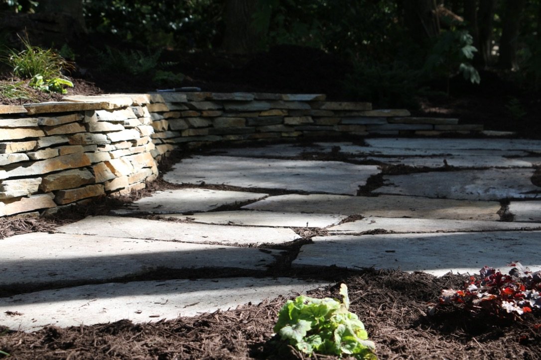 A stone pathway with cracked and uneven concrete slabs, bordered by a low stacked stone wall on the left, and surrounded by dark soil and small plants, with trees and foliage in the background.