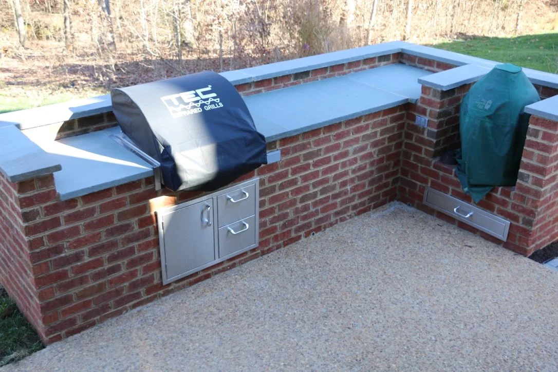Outdoor brick kitchen with two built-in grills, one covered with a black grill cover and the other with a green cover, set on a concrete patio with a wooded background.
