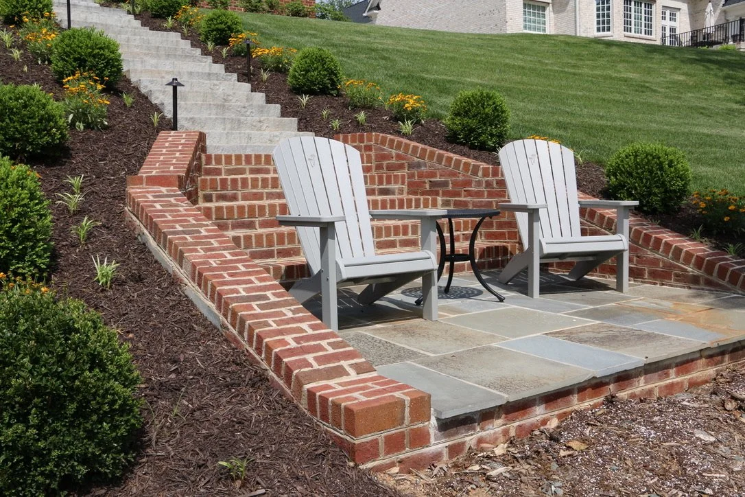 A small backyard patio with two gray Adirondack chairs, a round metal table, and brick retaining walls. Surrounding the patio are steps leading up a hill with landscaped bushes and flowers, with a green lawn and house in the background.
