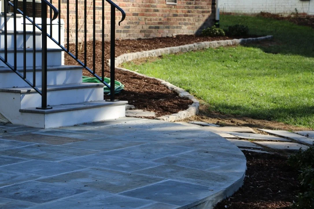Outdoor area showing a stone patio, stairs with black metal railings, new landscaping with soil and grass, and a brick house in the background.