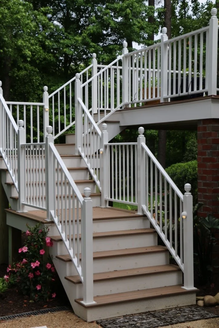 Outdoor white staircase with brown steps leading to a second-floor porch, surrounded by greenery and pink flowers at the bottom.
