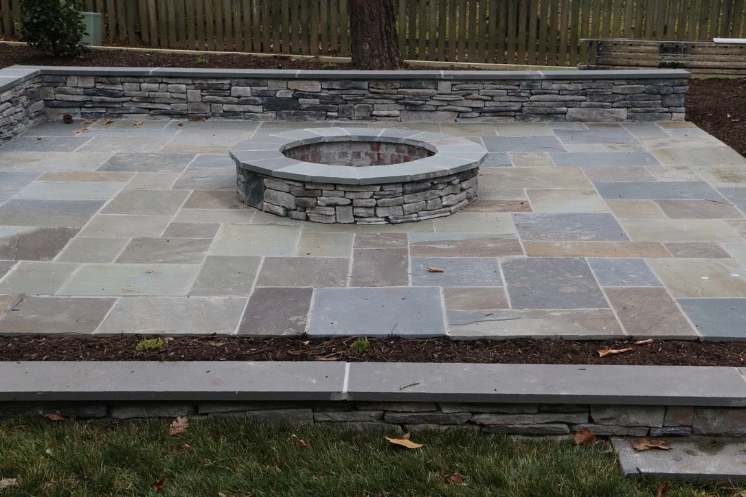 A stone patio with a built-in fire pit surrounded by a low stone wall, with grass and a wooden fence in the background.