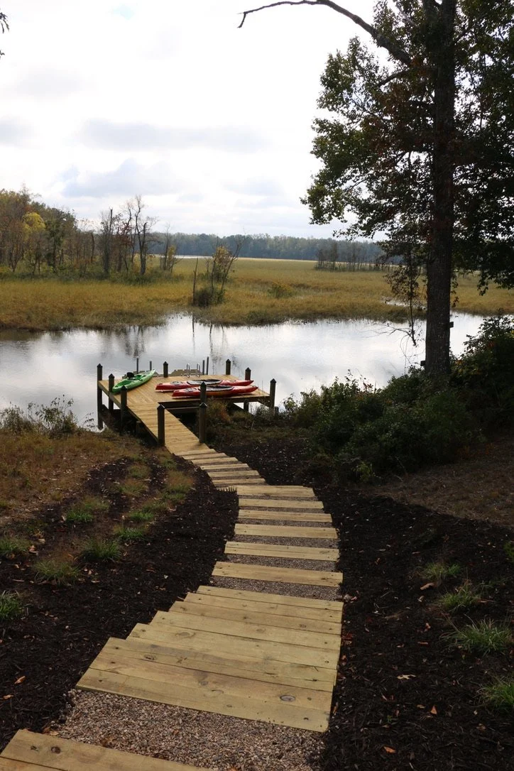 A wooden pathway leading to a small dock with several colorful kayaks on it, overlooking a calm lake surrounded by trees and open fields under a cloudy sky.