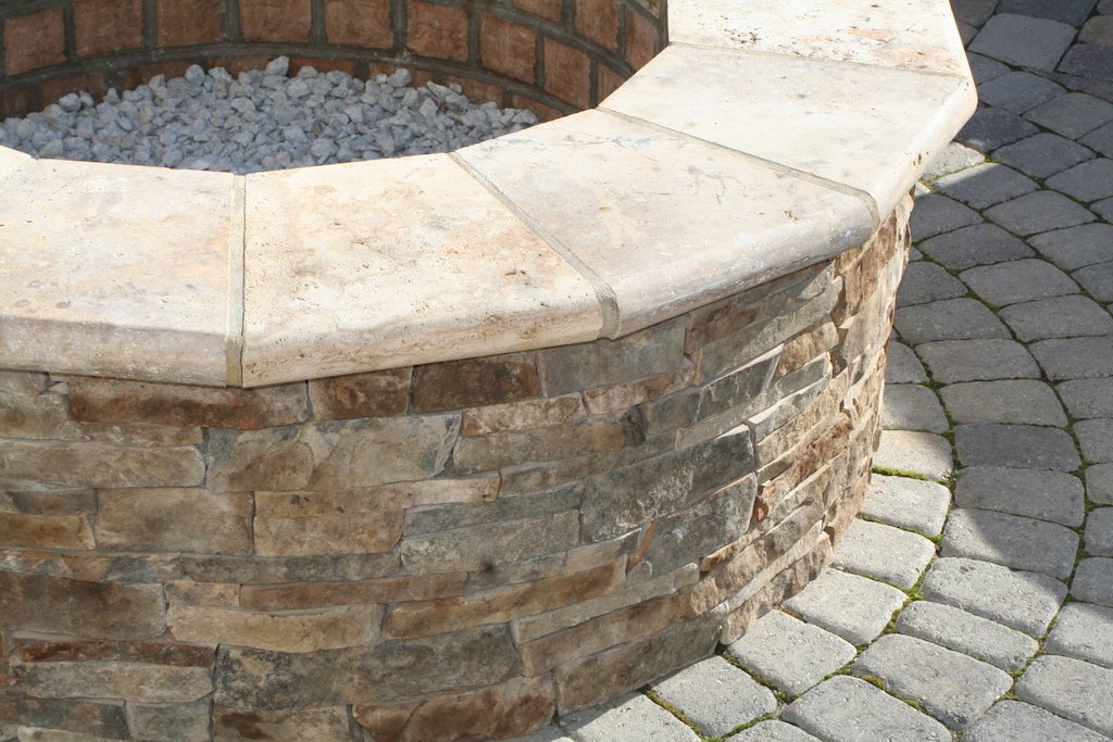 A close-up of a circular stone fire pit with a stone ledge, surrounded by gray cobblestone pavers.