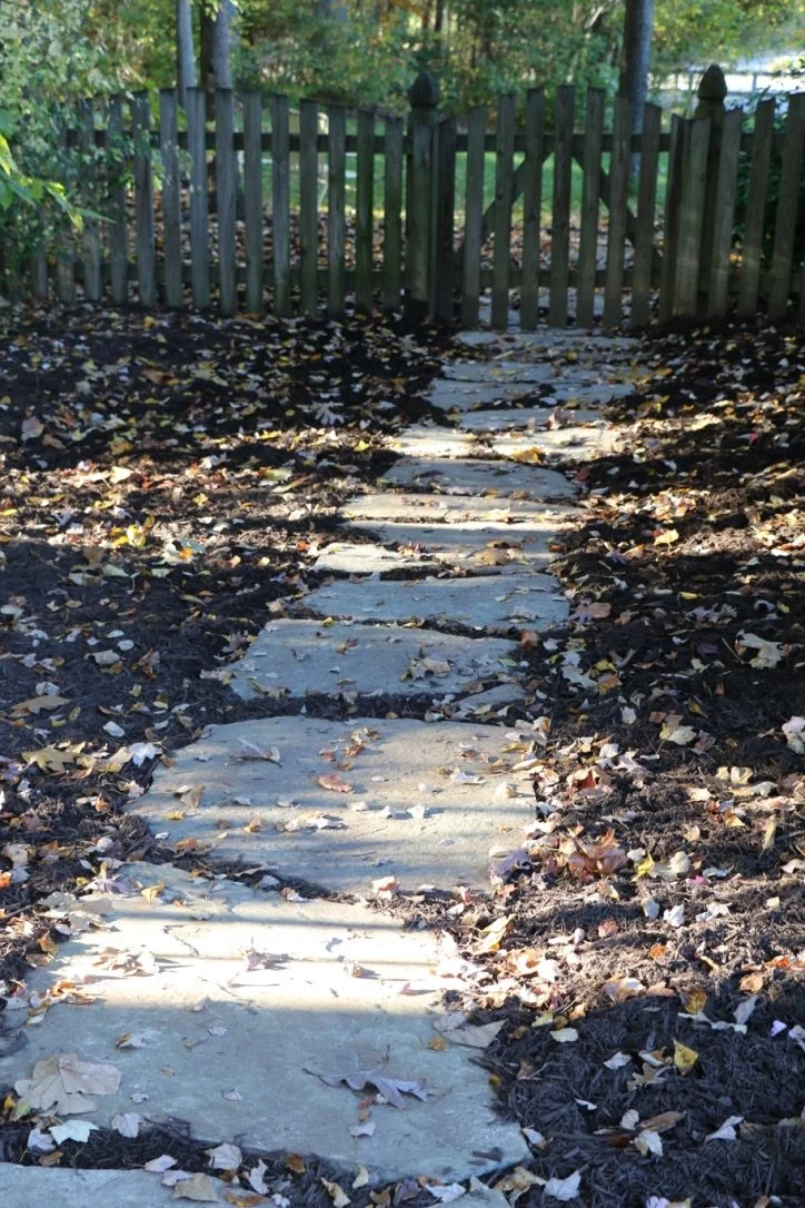 A stone pathway with fallen autumn leaves leading to a wooden fence in a backyard