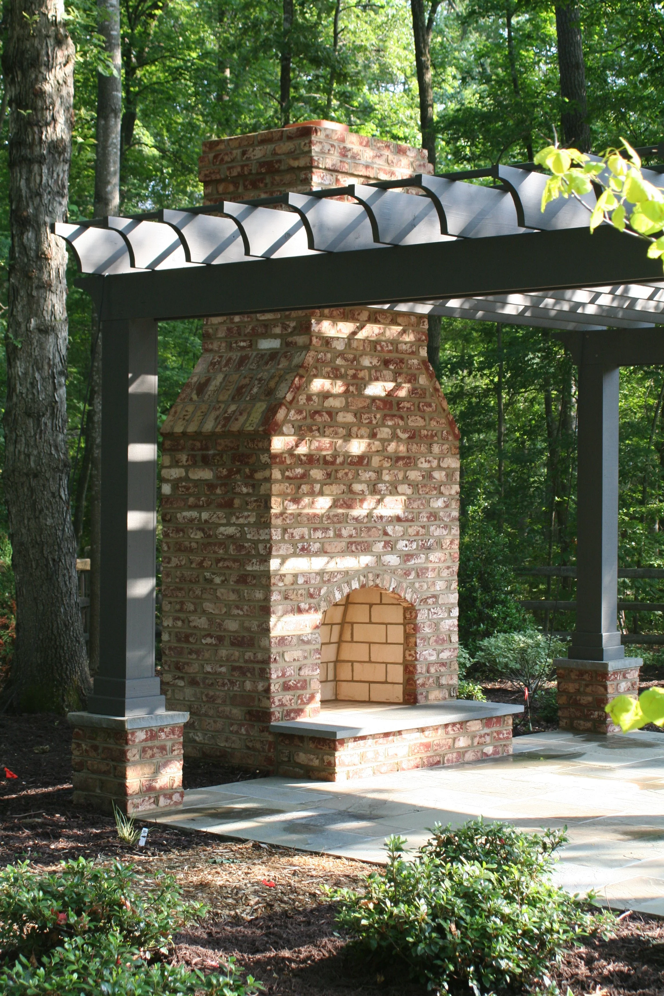 A brick outdoor fireplace with a chimney, set under a painted pergola in a wooded backyard.