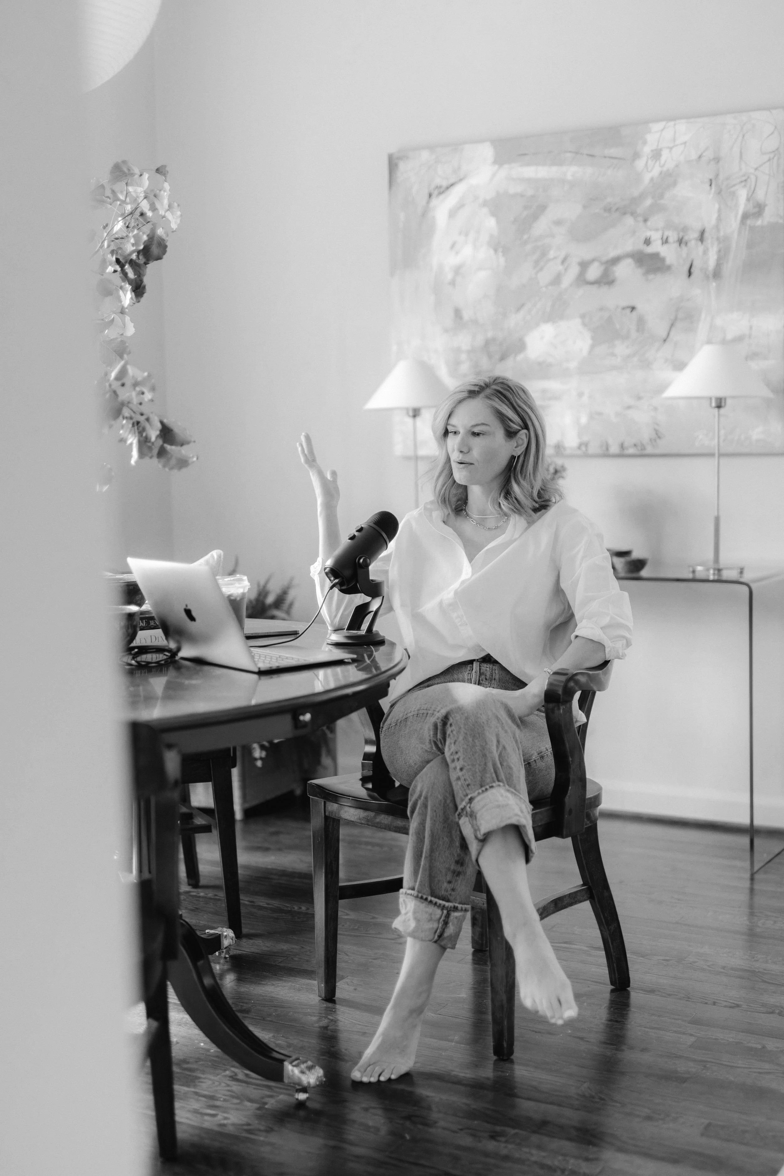 A woman with light wavy hair is sitting barefoot at a round table, speaking into a microphone, with a laptop in front of her and her hand raised. The room has a large abstract painting, two lamps, and a sideboard in the background.