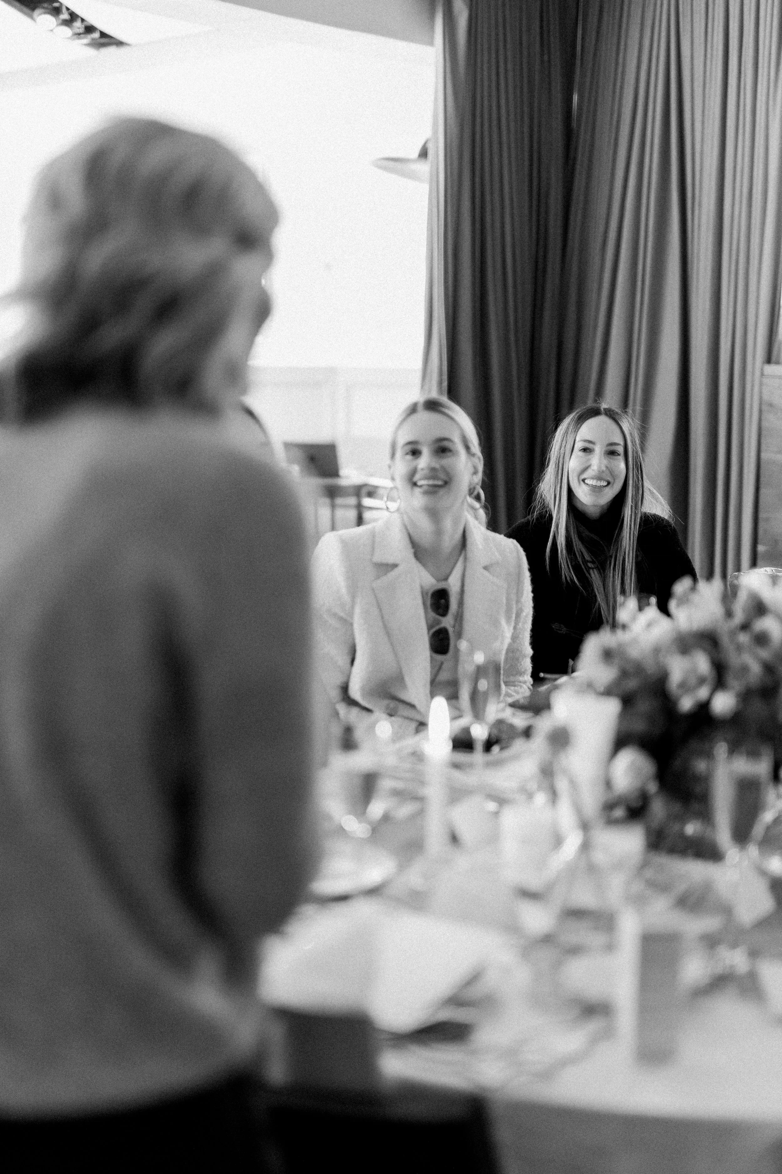 Three women sitting at a table during a social gathering or celebration, smiling and engaging in conversation. The image is in black and white.