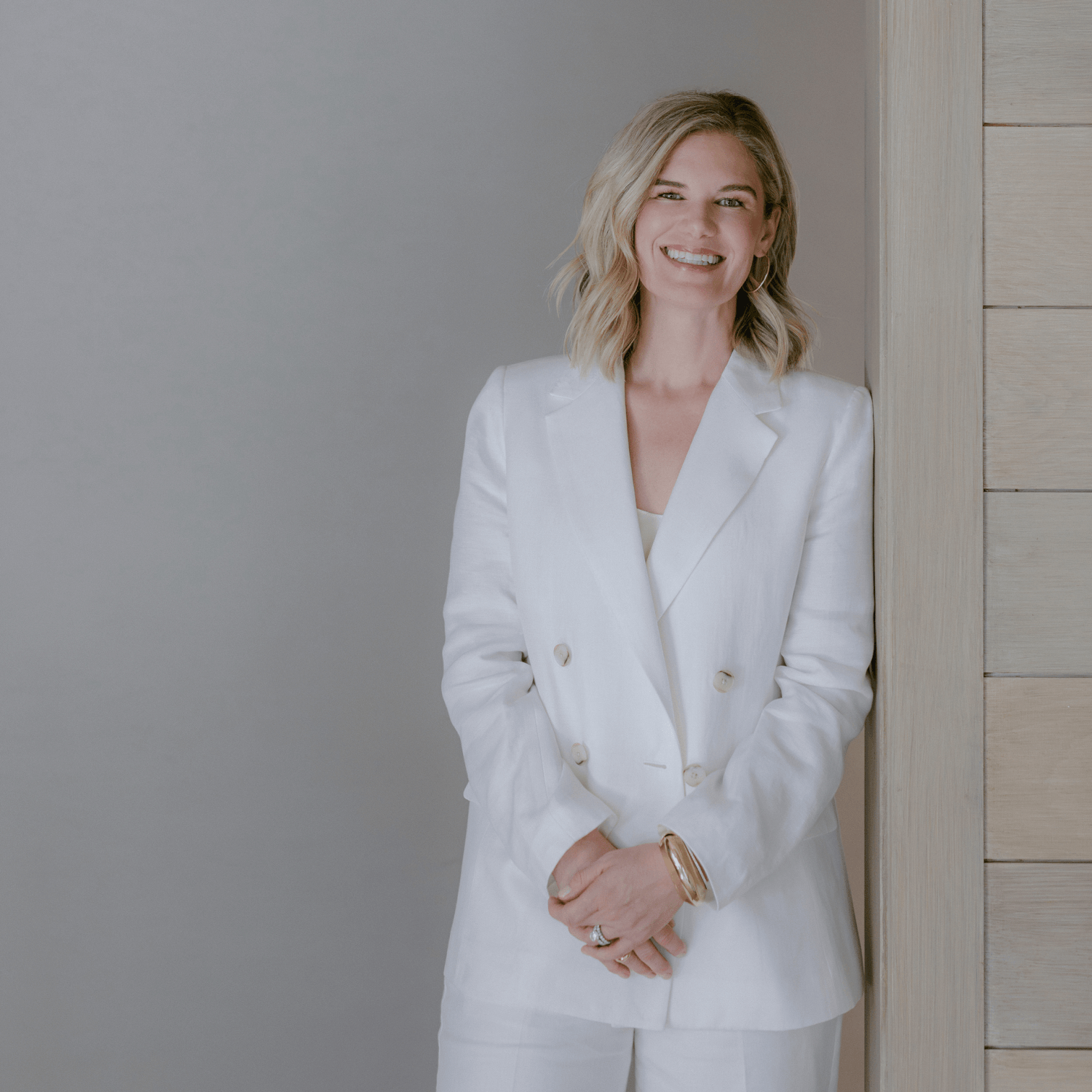 A woman in a white suit standing and smiling, leaning against a wooden wall.