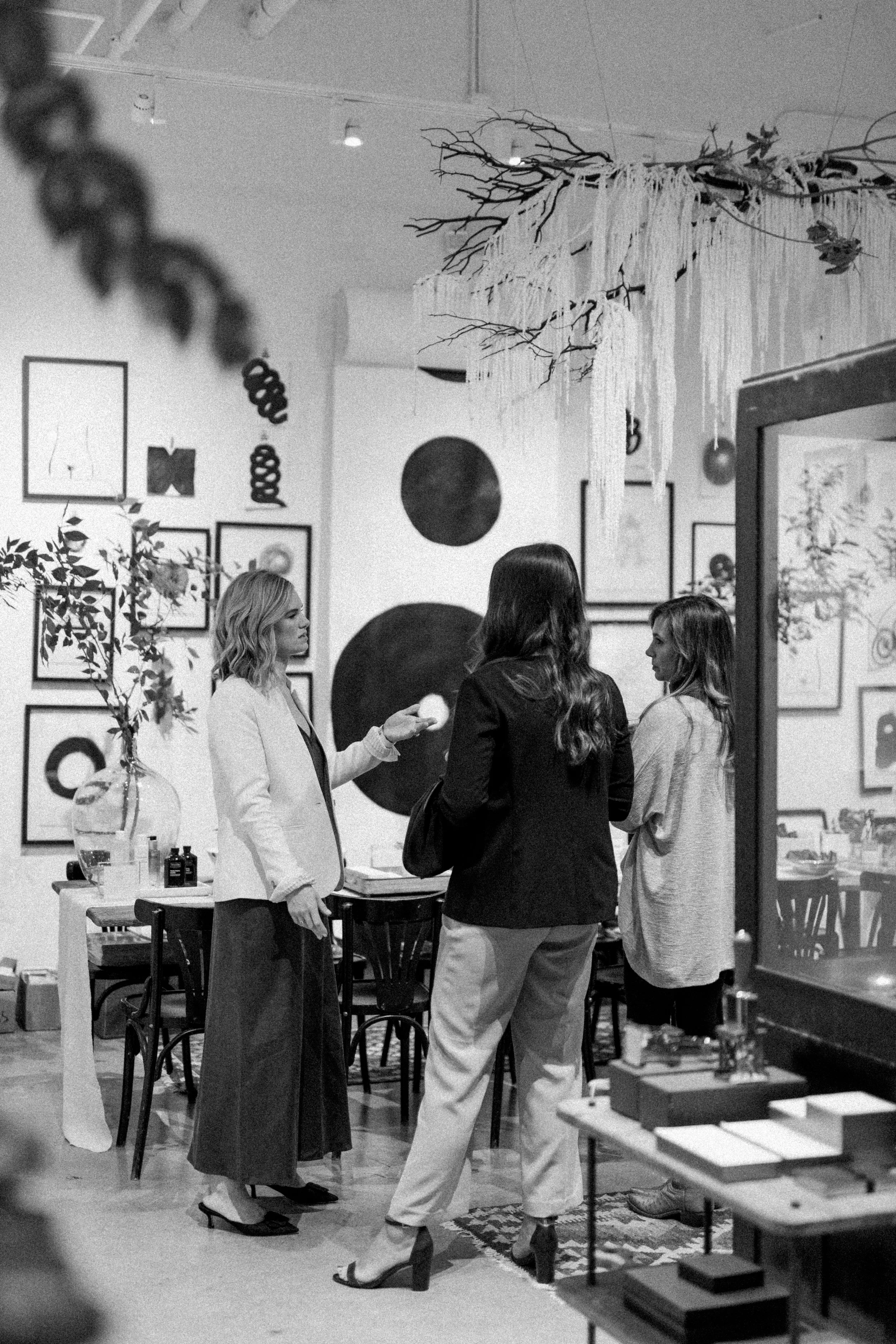 Three women standing and conversing in an art gallery with framed artworks on the wall and a decorative ceiling installation.