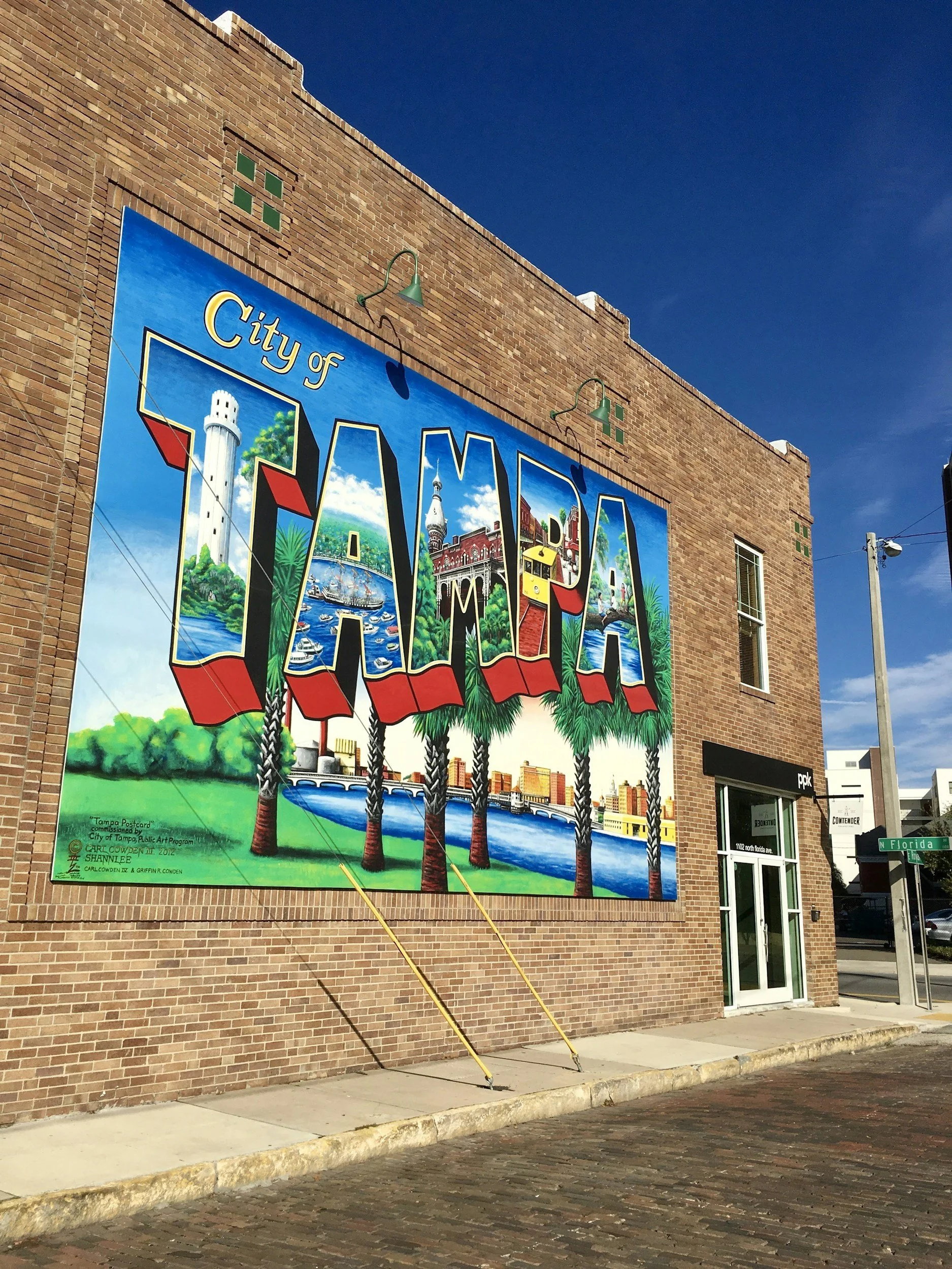 A mural on a brick building welcoming to the City of Tampa, featuring large colorful letters spelling 'TAMPA' with images of local landmarks, palm trees, and water.