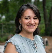 A woman with shoulder-length dark hair smiling outdoors in a park with trees in the background.