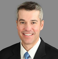 Headshot of a man with short gray hair, smiling, wearing a black suit, white shirt, and blue tie.