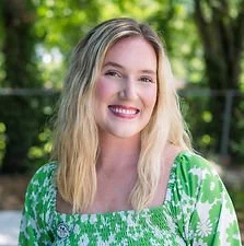 A smiling young woman with blonde hair wearing a green floral dress outdoors with trees in the background.